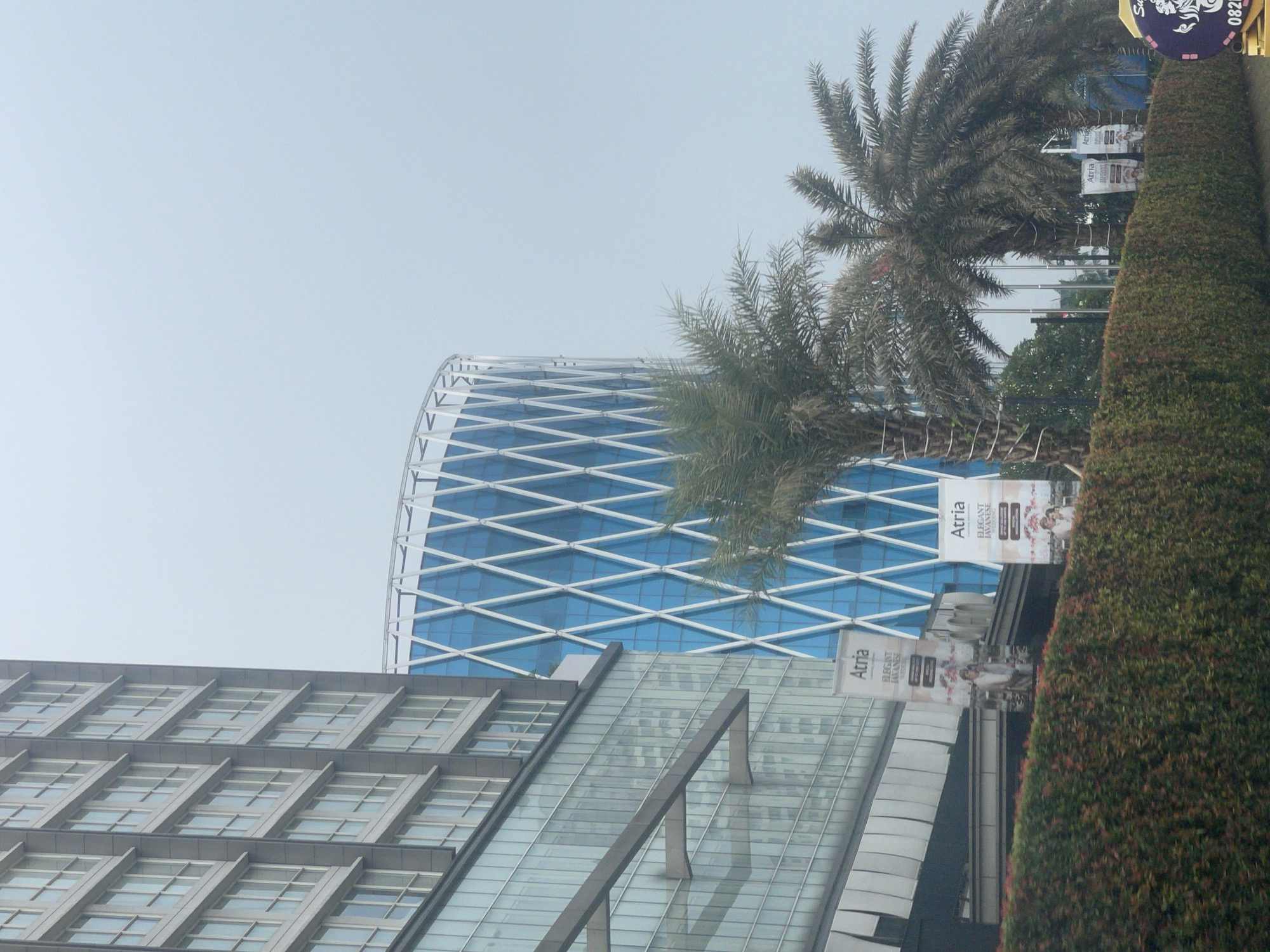 The image depicts a view of modern buildings and vegetation in Pakulonan Barat, Indonesia. On the left, a tall, rectangular building with a facade of many windows dominates the frame. Adjacent to it, to the right, is a distinctive cylindrical building with a blue glass exterior and a white diamond-shaped lattice pattern. In the foreground, a meticulously maintained hedge runs horizontally across the bottom of the image. Several palm trees are visible, with their fronds reaching upwards and obscuring parts of the buildings. Banners advertising "Atria Elegant Javanese" are placed in front of the hedge and near the buildings. The sky is overcast, indicating a cloudy day. The scene appears to be a street view, possibly from a vehicle, given the perspective and the presence of the hedge suggesting a roadside. There are no people clearly visible in the frame.