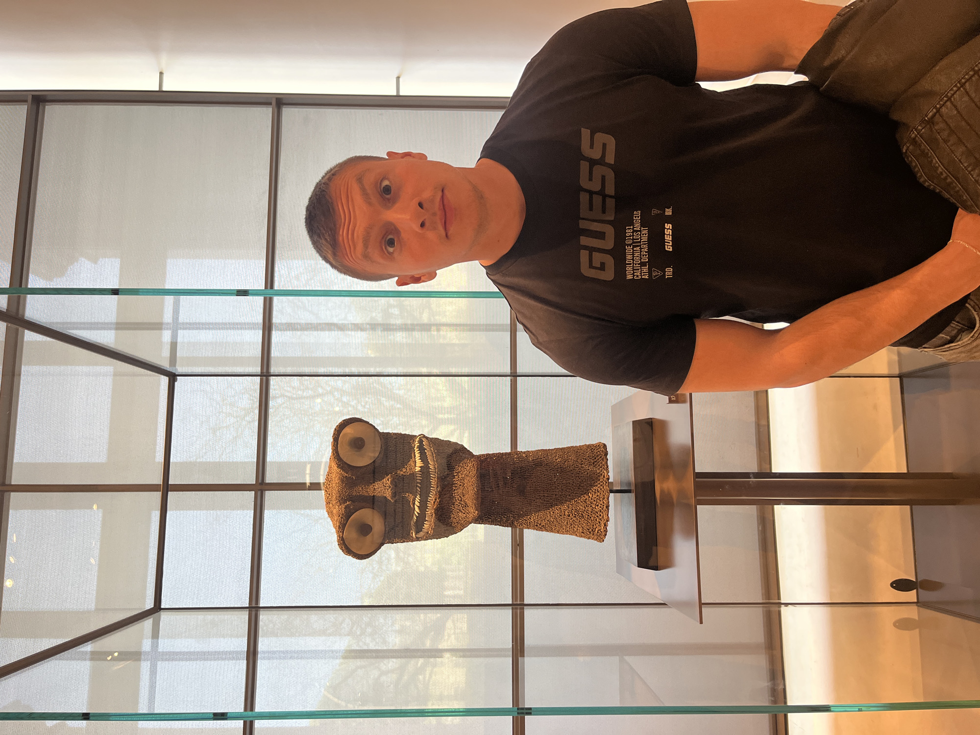 The image depicts a man posing in what appears to be a museum or art gallery in Paris, France. The man, with short, dark hair and fair skin, looks directly at the camera with a playful or surprised expression, featuring wide eyes and slightly raised eyebrows. He is wearing a black t-shirt that prominently displays the word "GUESS" in large white letters. Below this, smaller text reads "WORLDWIDE ©1981," "CALIFORNIA | LOS ANGELES," and "ATHL. DEPARTMENT," along with "TRD." and another "GUESS K."
Positioned to the man's right, inside a glass display case, is a distinctive exhibit: a brown, mask-like object. This artifact is intricately woven from a textured, possibly beaded or fiber material, and features large, light-colored, round eyes that protrude from its surface. It also has a wide, horizontal mouth with visible teeth-like elements. The object is mounted on a dark stand.
The background reveals a modern indoor setting with large windows composed of a grid of lighter panels, allowing ample daylight to illuminate the space. Reflections are visible on the glass surfaces of the display case and the windows, characteristic of a brightly lit exhibition hall during the daytime.