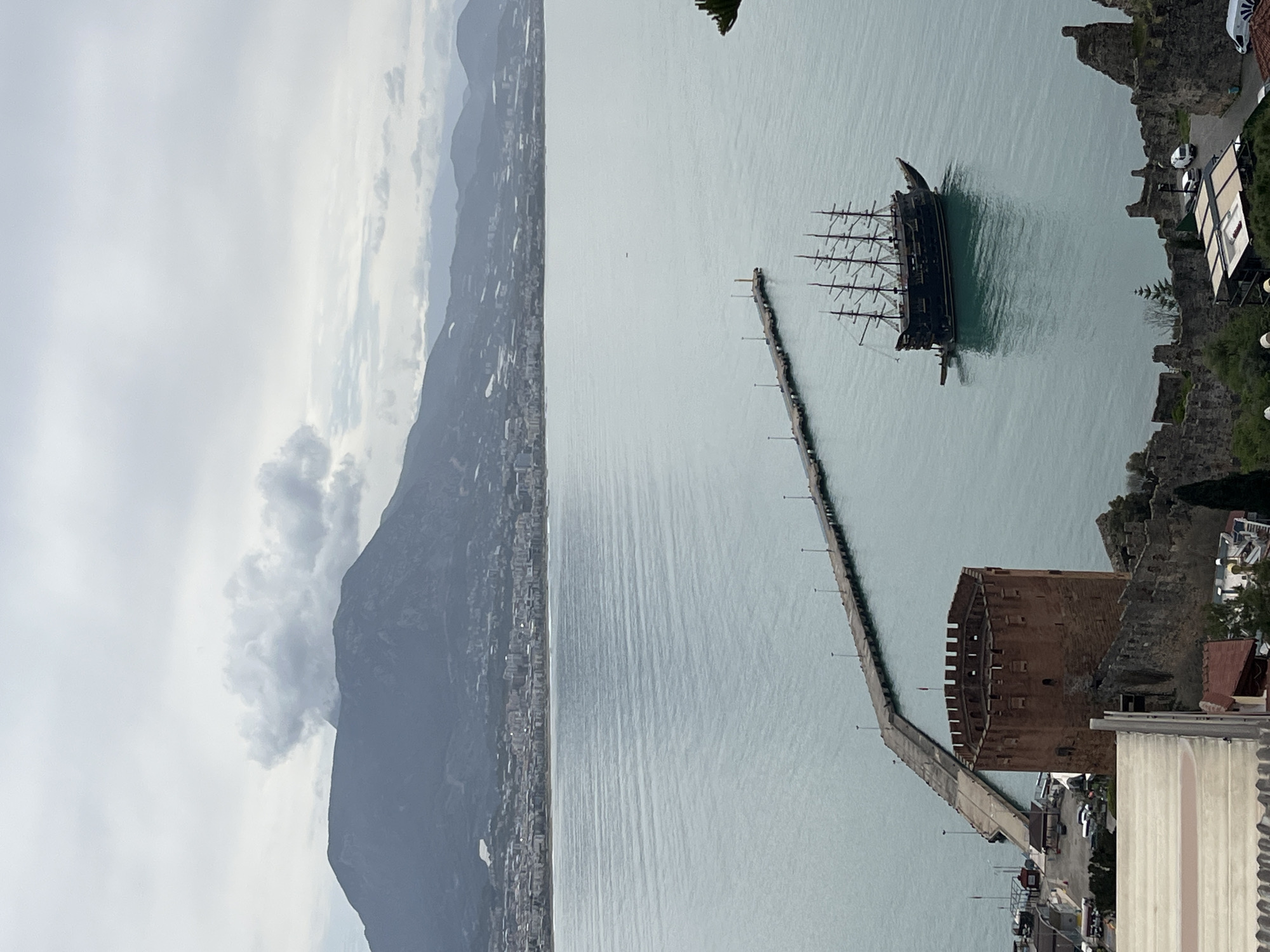 The image shows a high-angle view of Alanya, Turkey. A large, dark-colored pirate ship is docked near a long pier that extends into the water. At the base of the pier is the Red Tower, a historical fortress with visible battlements. Adjacent to the Red Tower are sections of old city walls and newer buildings. A coastal city stretches along the base of a tall mountain range. The weather appears overcast with cloudy skies.
