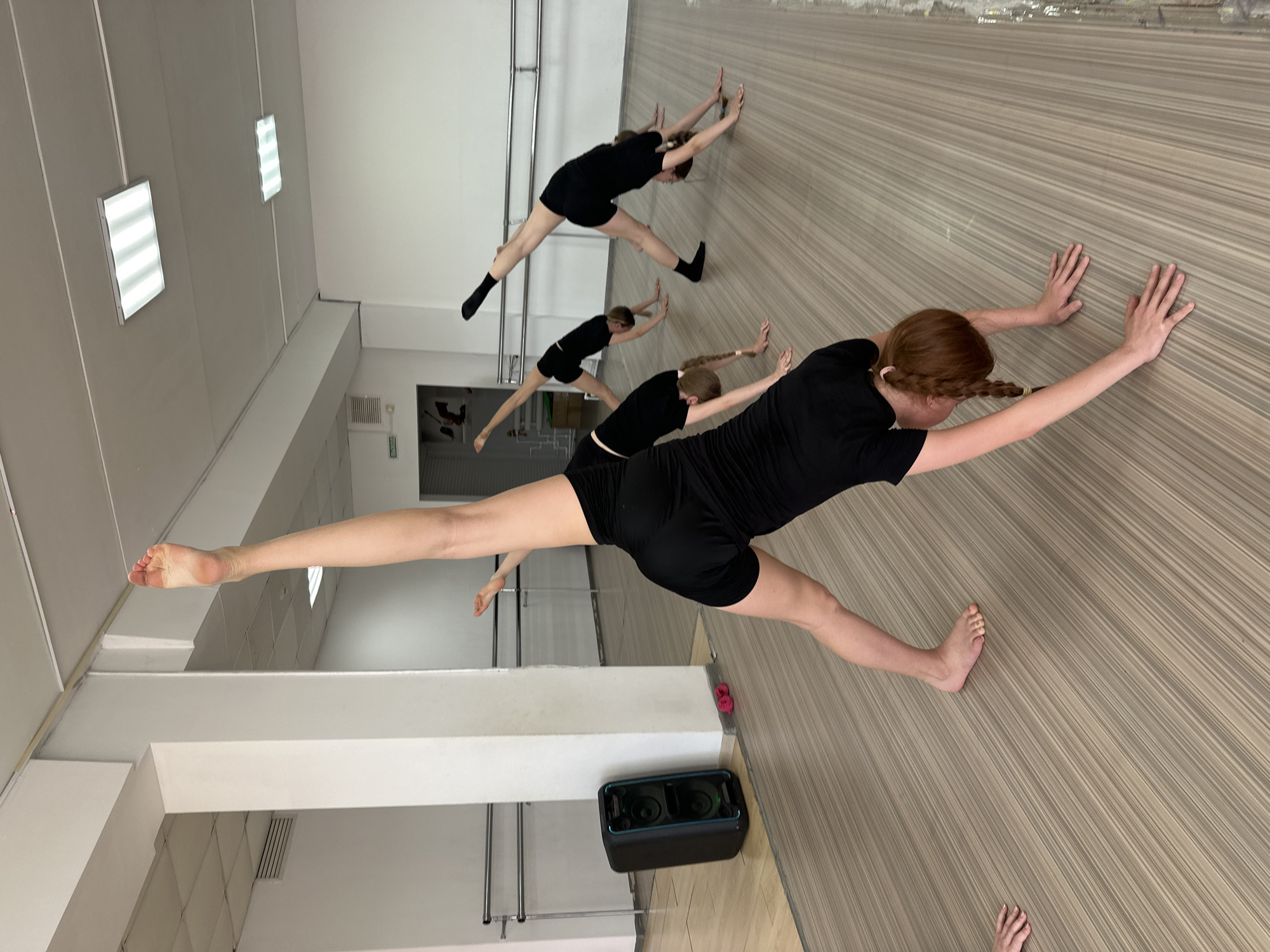 The image shows a dance studio in Votkinsk, Russia, where a group of female dancers are practicing. Five dancers in black leotards and shorts perform a stretching exercise, leaning forward against a wall with their legs in a straddle split. One dancer is in the foreground, closer to the camera, with red hair in braids. The studio has a light-colored wood-look floor and white walls. There is a large black speaker on the floor near the mirror. The lighting is provided by fluorescent ceiling fixtures. In the background, a doorway is visible.