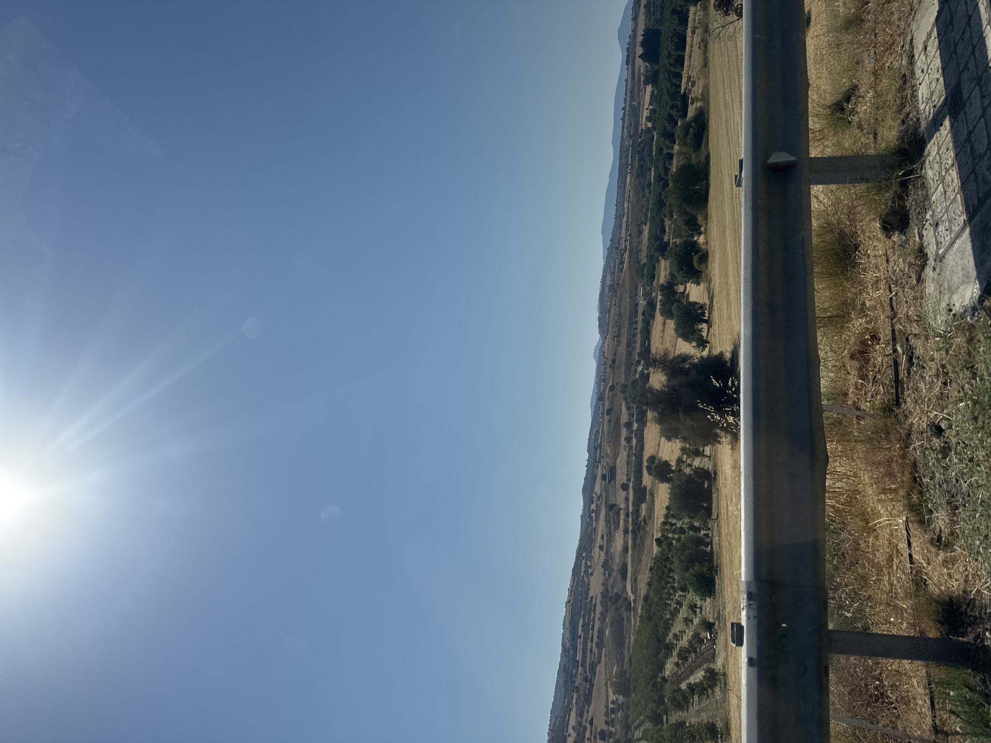 The image depicts a sun-drenched rural landscape under a clear blue sky. The sun's rays are strong, creating a lens flare effect. In the foreground, a weathered metal guardrail runs horizontally across the frame, supported by concrete posts. Beyond the guardrail, a field of dry, golden grass stretches towards a gently rolling terrain dotted with trees and crops. In the mid-ground, rows of young trees, possibly olive trees, are visible, suggesting agricultural activity. The background features a hazy horizon with distant hills and mountains. The overall scene suggests a dry, summery day in a rural area. The location is the vicinity of Chrysochou, Cyprus, indicated by the dry, Mediterranean vegetation and the characteristic landscape. No people or specific actions are discernible.