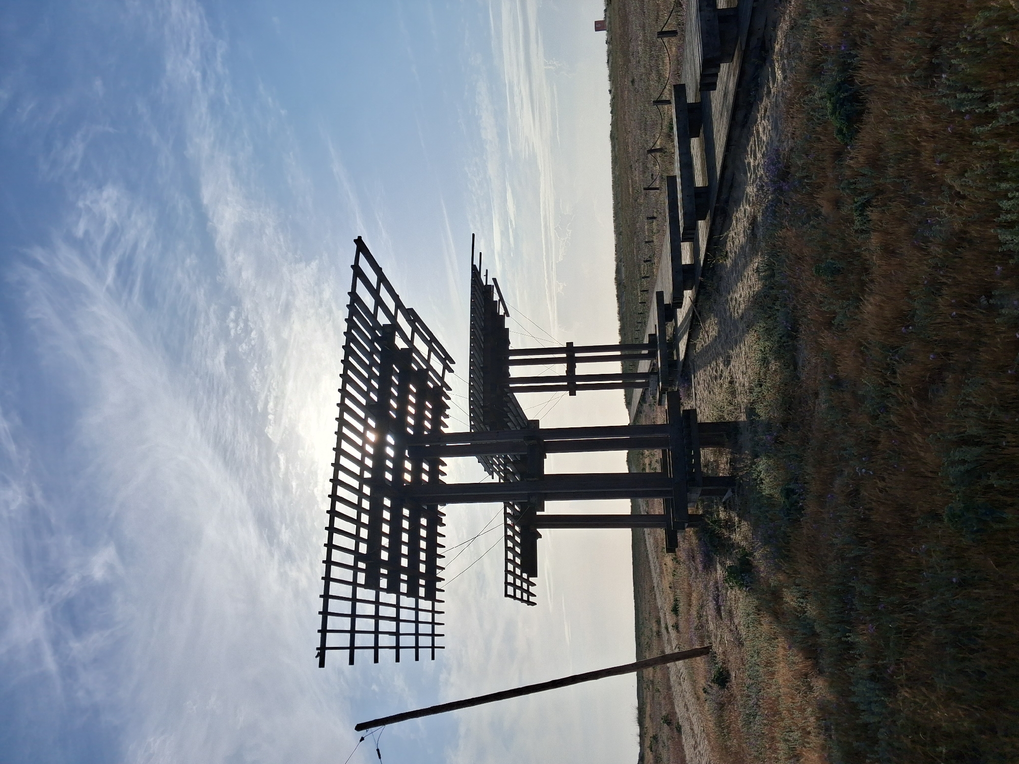 The image depicts a complex wooden structure against a bright sky with scattered clouds. The structure is composed of interconnected wooden grids and beams. The sun is shining through the grid, creating strong contrasts. The structure appears to be elevated and supported by a single pole. The surrounding environment consists of a field of dry grass and vegetation, suggesting a coastal or rural setting. A wooden walkway with railings is adjacent to the grassy area. Figueira da Foz, Portugal is not mentioned in the photo.