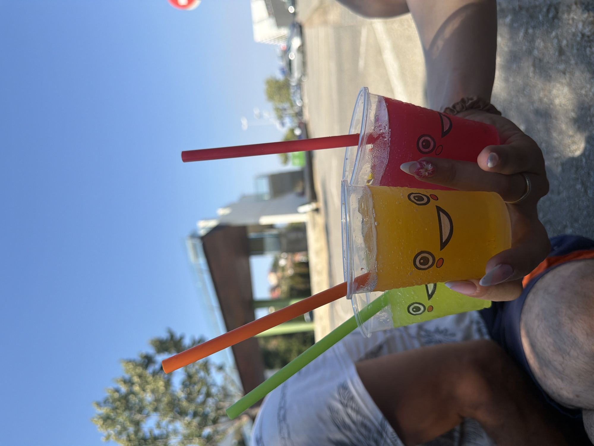 In Santa Susanna, Spain, a close-up shot captures three plastic cups with cartoon faces filled with brightly colored beverages and straws. The cup on the left contains a lime-green drink, the middle cup a vibrant orange drink, and the right cup a deep pink beverage. The cups are held by a person whose hand, with meticulously manicured nails, is visible in the foreground. The background is slightly blurred, showing a clear blue sky, trees, and a building structure, likely an outdoor recreational area or a part of the resort infrastructure. The scene appears to be outdoors on a sunny day, with some shadows visible on the ground, suggesting it's daytime.