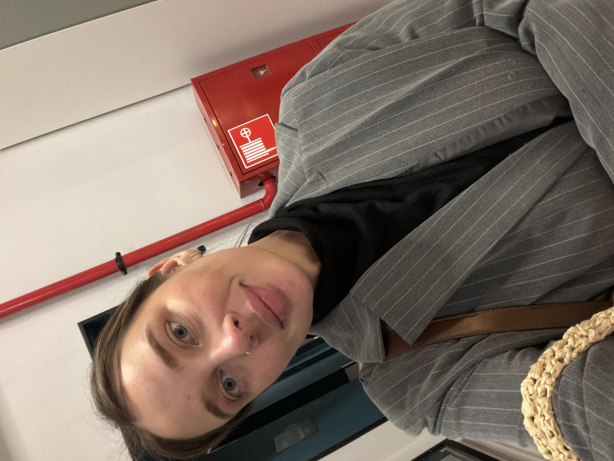 In a selfie, a woman with light brown hair pulled back, blue eyes, and a nose ring smiles at the camera. She is wearing a grey pinstripe blazer over a black turtleneck and a brown strap is visible over her shoulder. To her right, a red fire alarm cabinet is mounted on a pale wall, with a red pipe extending from the top of the cabinet upwards. A portion of a wicker bag is visible in the lower left corner. The scene appears to be indoors, potentially in a public building in Moscow, Russia.