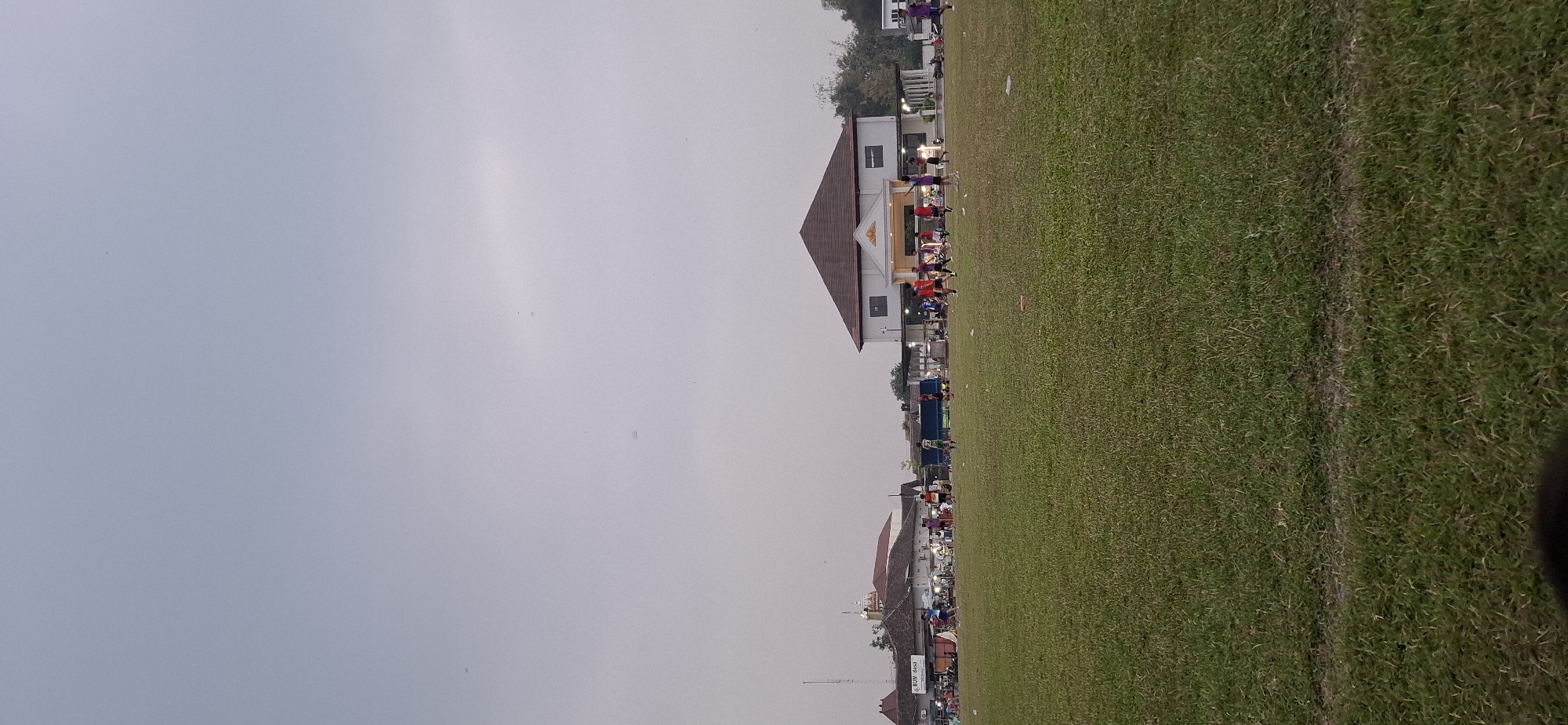 The image depicts a soccer field in Wonorejo, Indonesia, under a cloudy sky. A group of people, likely boys or young men, are playing soccer on the field, some in red jerseys and others in darker colors. In the background, there are several buildings, including a white building with a brown, pointed roof, and other structures that appear to be shops or market stalls. The field is covered in green grass, with some scattered debris. The scene appears to be an outdoor recreational area.