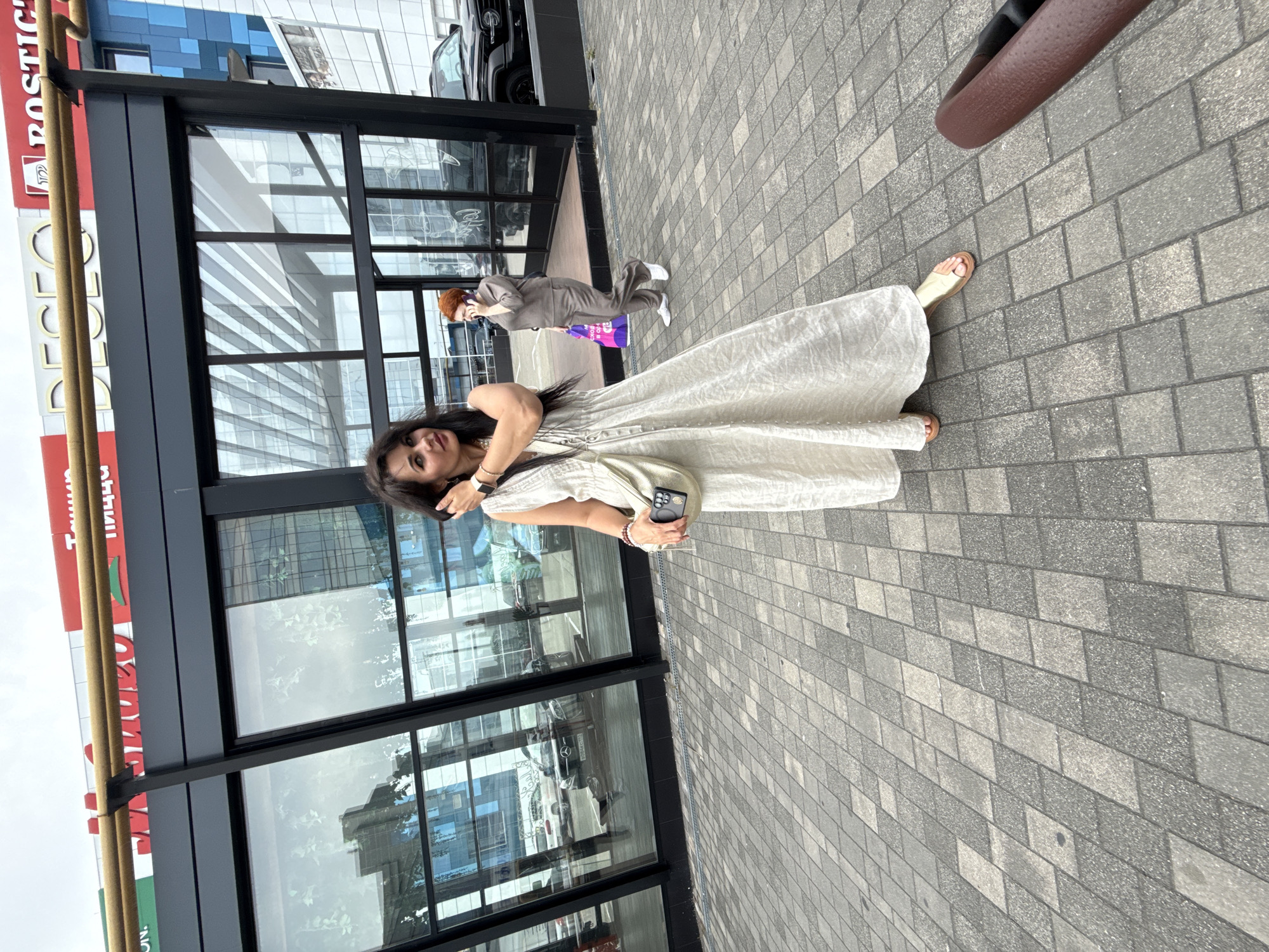 A woman in a long, cream-colored dress and gold sandals stands on a patterned pavement outside a modern building with large glass windows. She is holding her phone in her left hand and touching her hair with her right. Behind her, another woman in a brown jumpsuit is on the phone. The building has a red and gold sign above it, with the word "SOCHI" visible. The sky is overcast.
