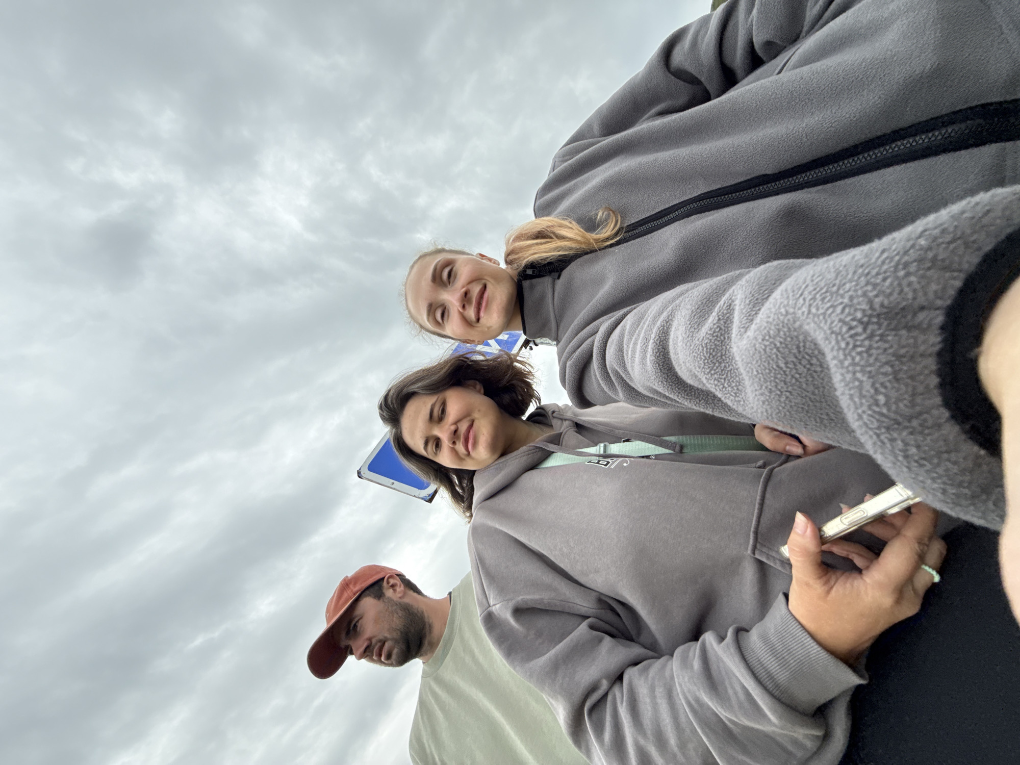 A low-angle, wide shot captures three individuals standing outdoors under a cloudy sky. On the left, a man wearing a light green t-shirt and an orange baseball cap looks upwards and to the left, his face partially obscured by the cap. In the center and foreground, a woman with dark, shoulder-length hair smiles towards the camera. She is wearing a gray hooded sweatshirt and has a mint-colored strap visible around her neck. Her left hand holds a silver object, possibly a phone. To her right, another woman with blonde hair pulled back in a ponytail also smiles at the camera. She is wearing a gray fleece jacket with a black zipper. A blue and white sign is visible behind the central woman. The image appears to be taken in Teriberka, Russia.