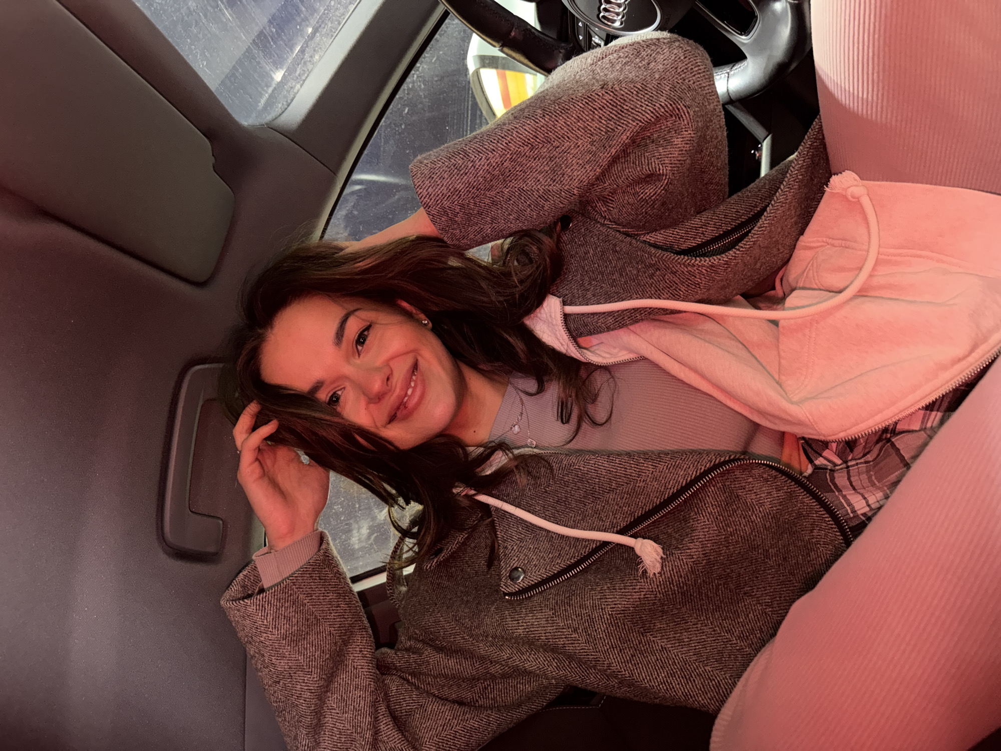 A young woman with long, dark brown hair smiles brightly at the camera while reclining in the passenger seat of an Audi car, located in Krasnoyarsk, Russia. She is wearing a grey herringbone patterned jacket over a light pink hoodie and a light grey long-sleeved top. Her expression is cheerful and relaxed. The photograph is taken from a high angle, looking down at her.
The car's interior is visible, including a dark sun visor and the "AUDI" logo clearly displayed on the steering wheel. The lighting appears dim with a warm, reddish-pink hue on the right side of the frame, suggesting an evening or nighttime setting. Through the car windows, indistinct blueish-grey structures are visible, possibly indicating an urban environment or a parking garage. The woman's right hand is casually placed in her hair above her head.