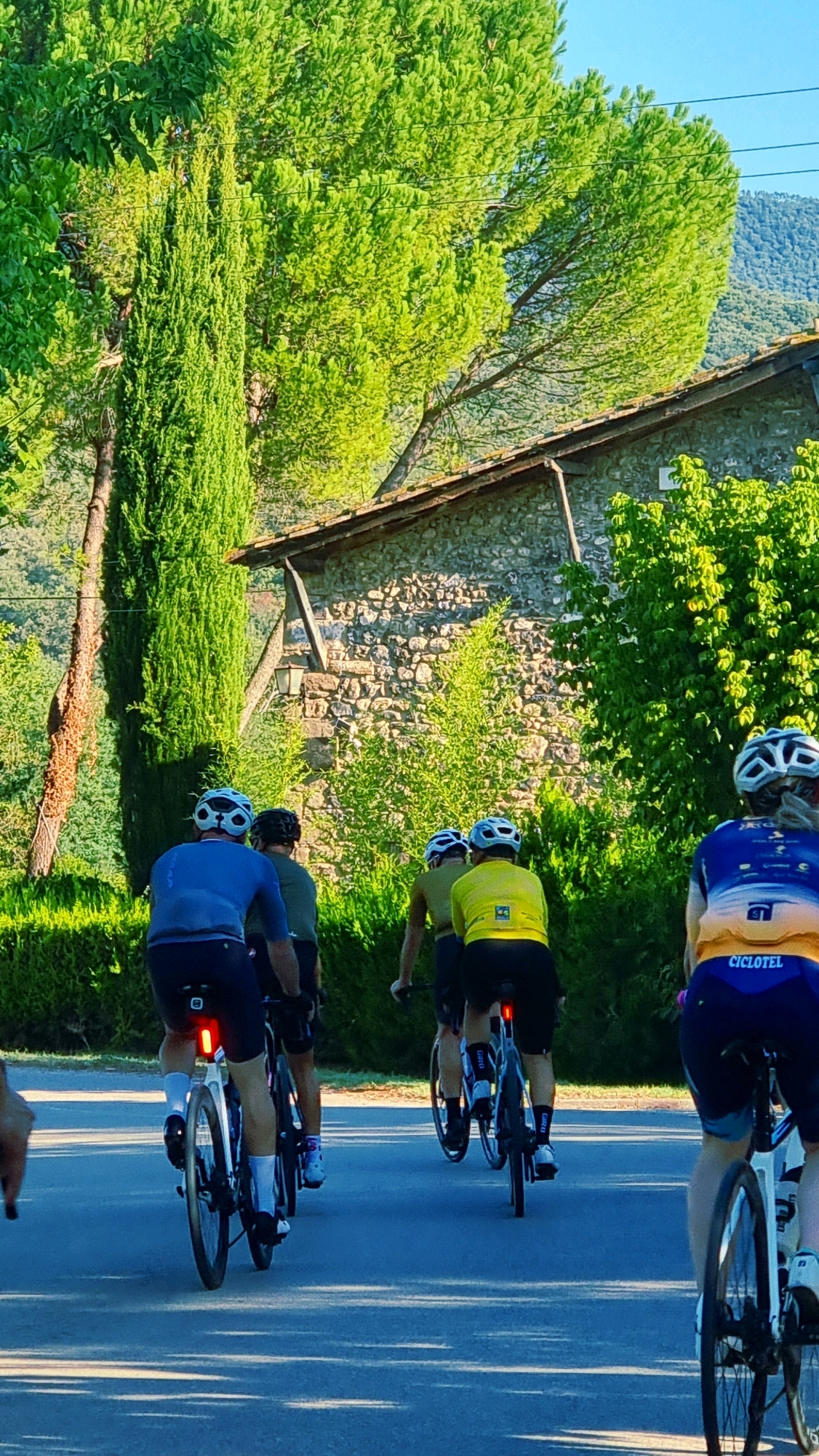 In a sun-drenched scene, four cyclists, seen from behind and facing away from the camera, pedal along a paved road. They are equipped with helmets and cycling attire. The cyclist in the foreground on the right wears a blue and yellow jersey with "CICLOTEL" visible. The cyclist in the middle foreground wears a yellow jersey and black shorts, with a red rear light on their bicycle. To their left, another cyclist in a blue jersey and black shorts is also visible. A fourth cyclist, partially obscured, is to the far left. The environment features lush green trees, a stone building with a tiled roof, and a distant hillside. The overall impression is a picturesque, sunny cycling excursion.