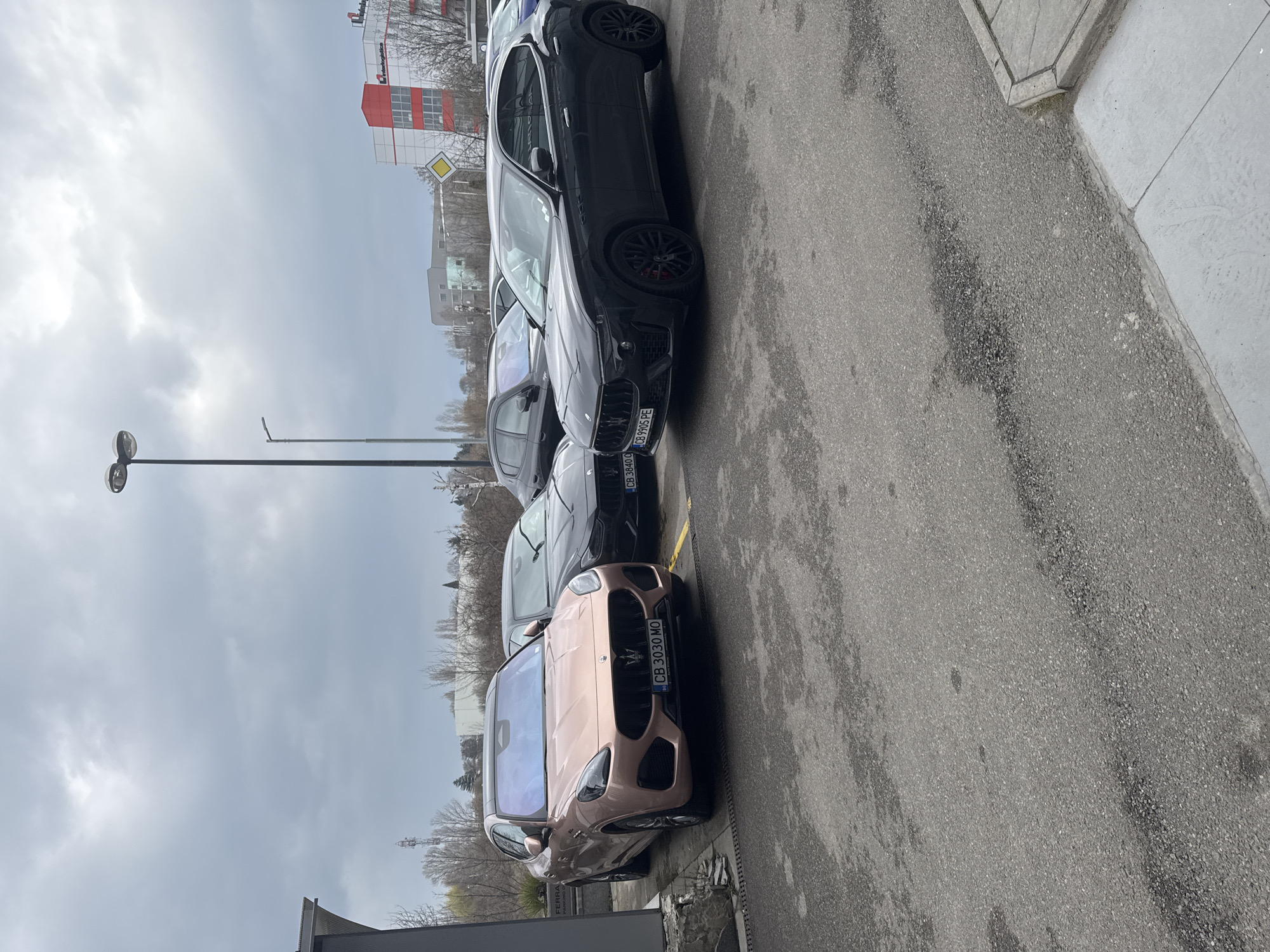 The scene depicts an outdoor parking lot at the FERRA Motors Maserati dealership on blvd. Tsarigradsko shose 166, Sofia, Bulgaria, under an overcast sky. Three luxury vehicles are prominently displayed. In the foreground, a rose gold (or copper-colored) Maserati Grecale SUV, license plate CB 3030 MO, is parked at an angle, its front left wheel slightly turned. Immediately behind it, a black Maserati Grecale SUV is partially visible, bearing a license plate starting with CB 3840. To the right, a black Maserati Levante SUV, license plate CB 9905 PE, is parked, showcasing black multi-spoke wheels and red brake calipers. Additional dark-colored vehicles are visible further in the background.
The ground consists of gray asphalt pavement with varying shades and dark, irregular stains or wet patches. A concrete curb and sidewalk section are visible on the far right. A dark-colored building with "FERRA PARKING" text partially visible on a sign is to the immediate left of the rose gold vehicle. In the mid-ground and background, deciduous trees and shrubs are bare, indicative of late winter or early spring. Beyond the trees, various commercial and industrial buildings are present. A prominent multi-story building with a white facade and distinct red rectangular sections, displaying the "Interlogistica" logo, is visible in the right background, along with a yellow rhombus-shaped priority road sign. Two street lamps are positioned in the mid-ground, one featuring dual light fixtures. A communication tower stands on the distant horizon line. No people or active interactions are observed in the image.