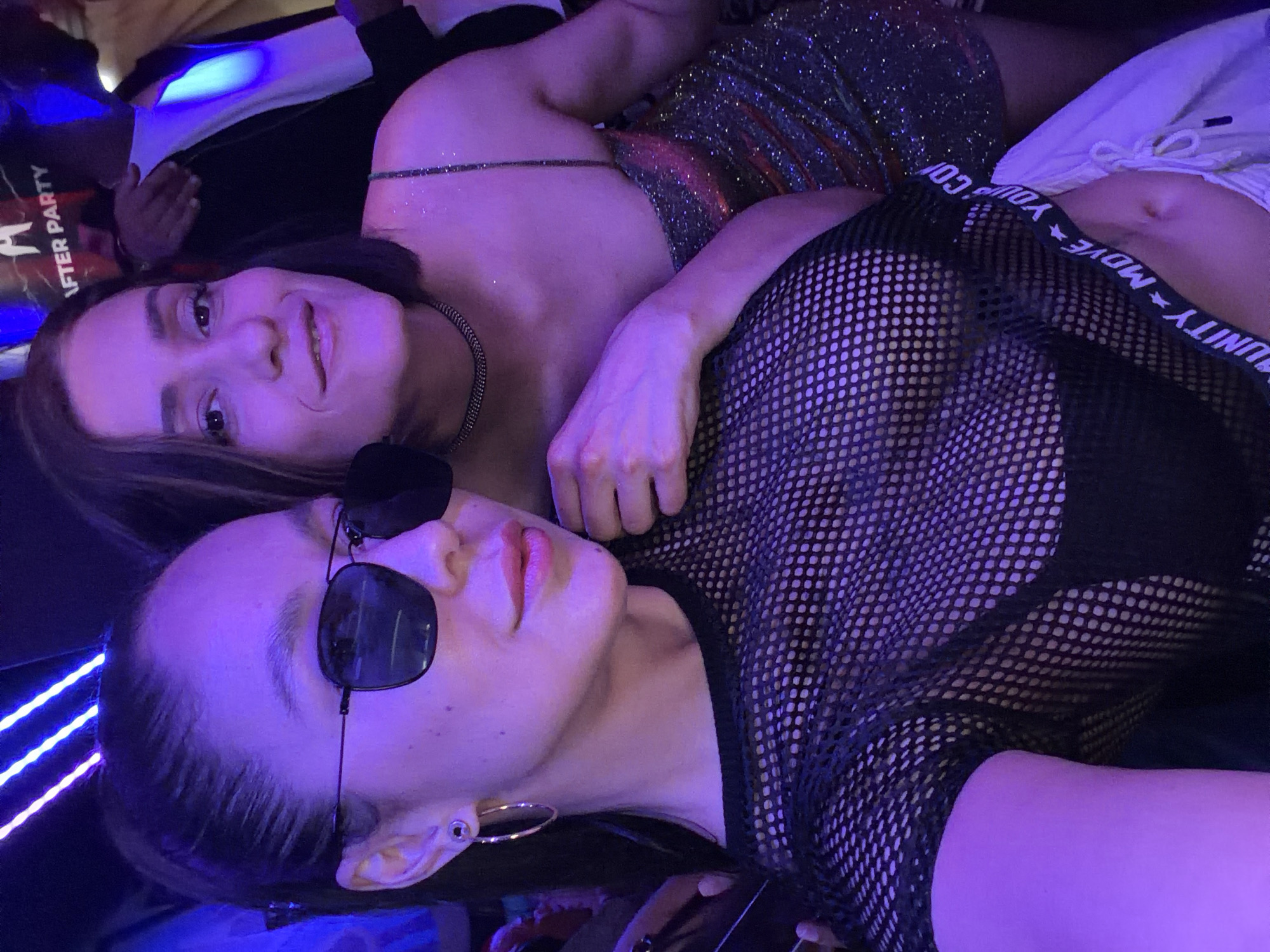 Two women are lying down next to each other in a club, illuminated by blue and purple lighting. One woman is wearing a sparkly dress and a chain necklace. She is looking at the camera and smiling. The other woman is wearing a black mesh top with a sports bra underneath and sunglasses. She is looking to the side. The scene is set in an "After Party". The location in the city of Barcelona, Spain cannot be determined from the image.