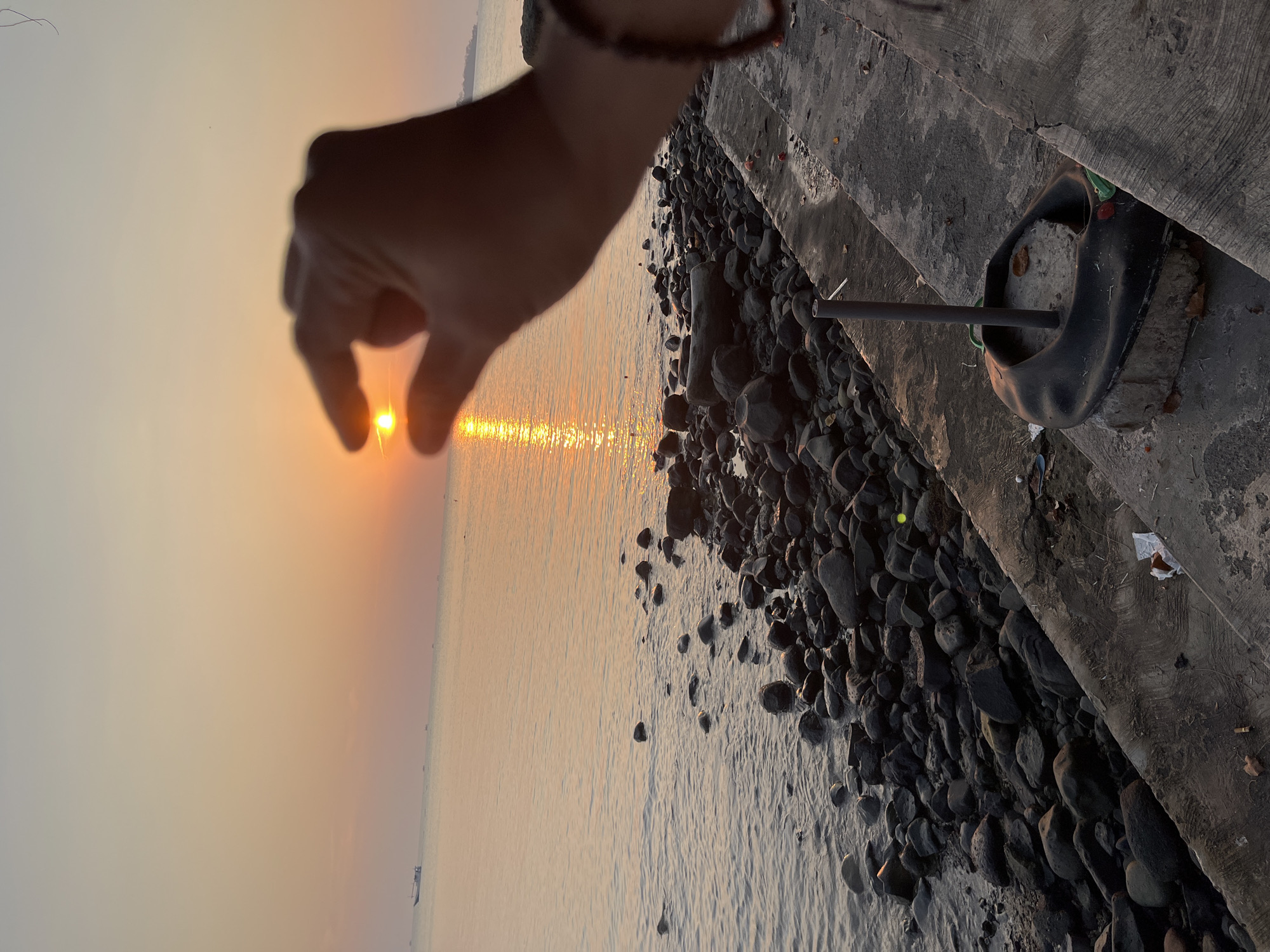 The image shows a hand holding the sun between its fingers at sunset, with the ocean in the background. The sun appears as a small, bright orb of light. The ocean is calm, reflecting the orange and yellow hues of the sunset. The foreground features a rocky shoreline with a concrete embankment. The scene is likely located in Jepara, Indonesia, a coastal city known for its beaches and maritime activities. The overall impression is one of a peaceful and picturesque sunset, with the hand's gesture adding a sense of wonder and scale to the natural spectacle.
