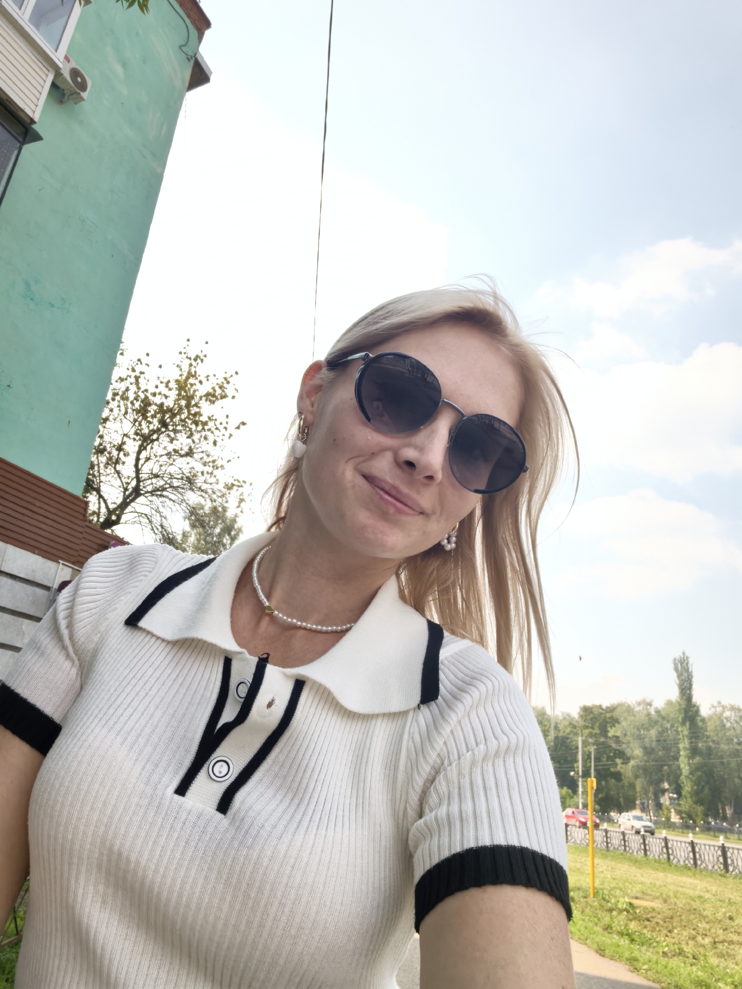 A blonde woman in her 30s with light skin and straight hair is taking a selfie outdoors. She is wearing round, dark sunglasses, a white short-sleeved collared shirt with black trim, and a pearl necklace with a small gold pendant. She has pearl earrings. The background shows a light blue sky with some clouds, a portion of a green building on the left, a tree, and a street with cars and trees in the distance. The image appears to have been taken in Sterlitamak, Russia, given the context provided.