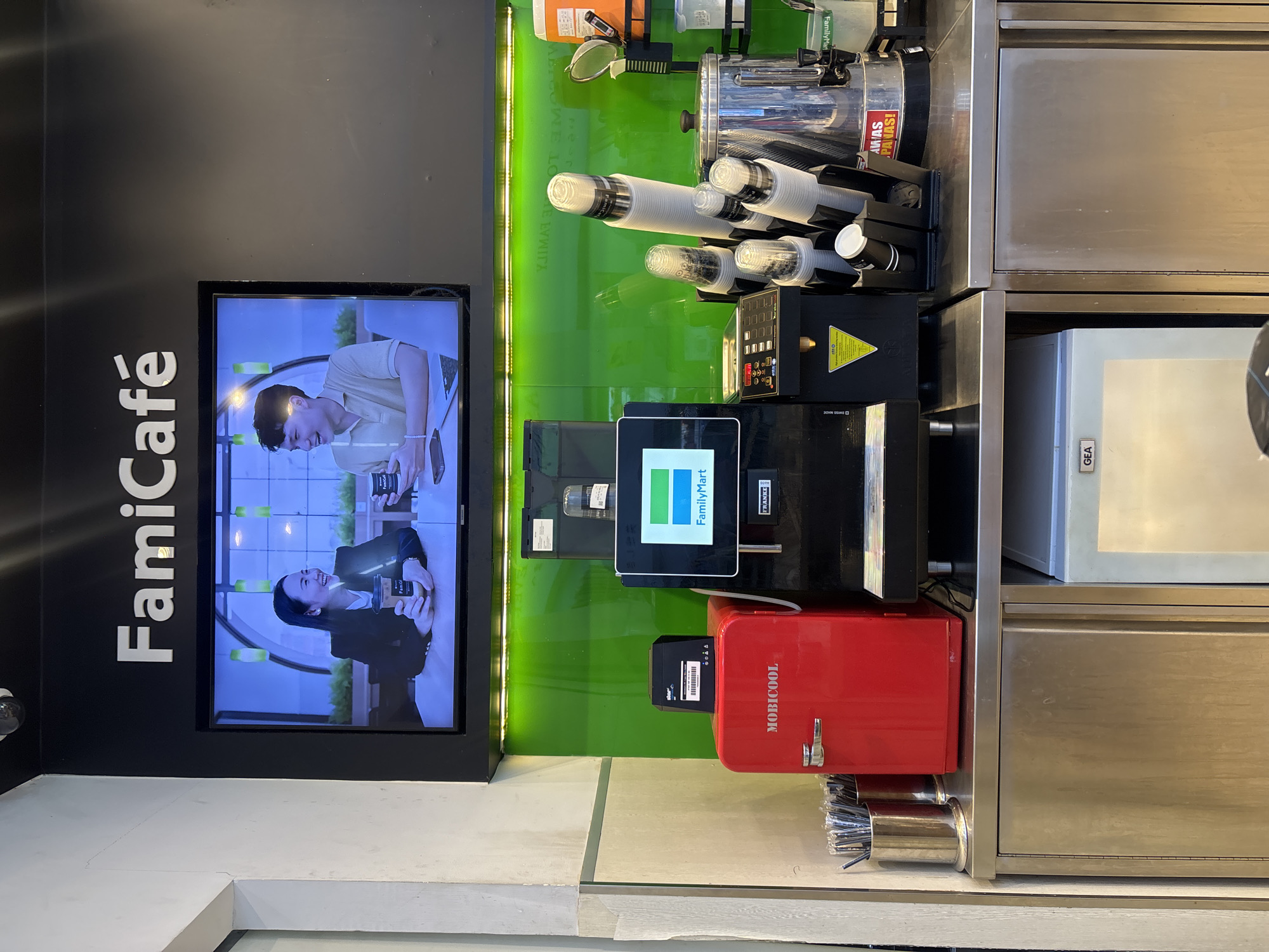 The image shows a FamiCafe counter display area in West Jakarta, Indonesia. A large screen TV displays a commercial for the cafe featuring two individuals enjoying drinks. Below the TV, a green tiled backsplash is visible. On the counter, a red Mobicoool mini-refrigerator is present, along with various coffee-making equipment, including a coffee machine with a FamilyMart logo, stacks of cups, and a large metallic dispenser. To the left, a cylindrical metal container holds numerous straws. In the lower portion of the image, stainless steel cabinets are visible, one of which houses a GEA branded appliance. The overall scene suggests a functional beverage service area within a FamiMart convenience store.