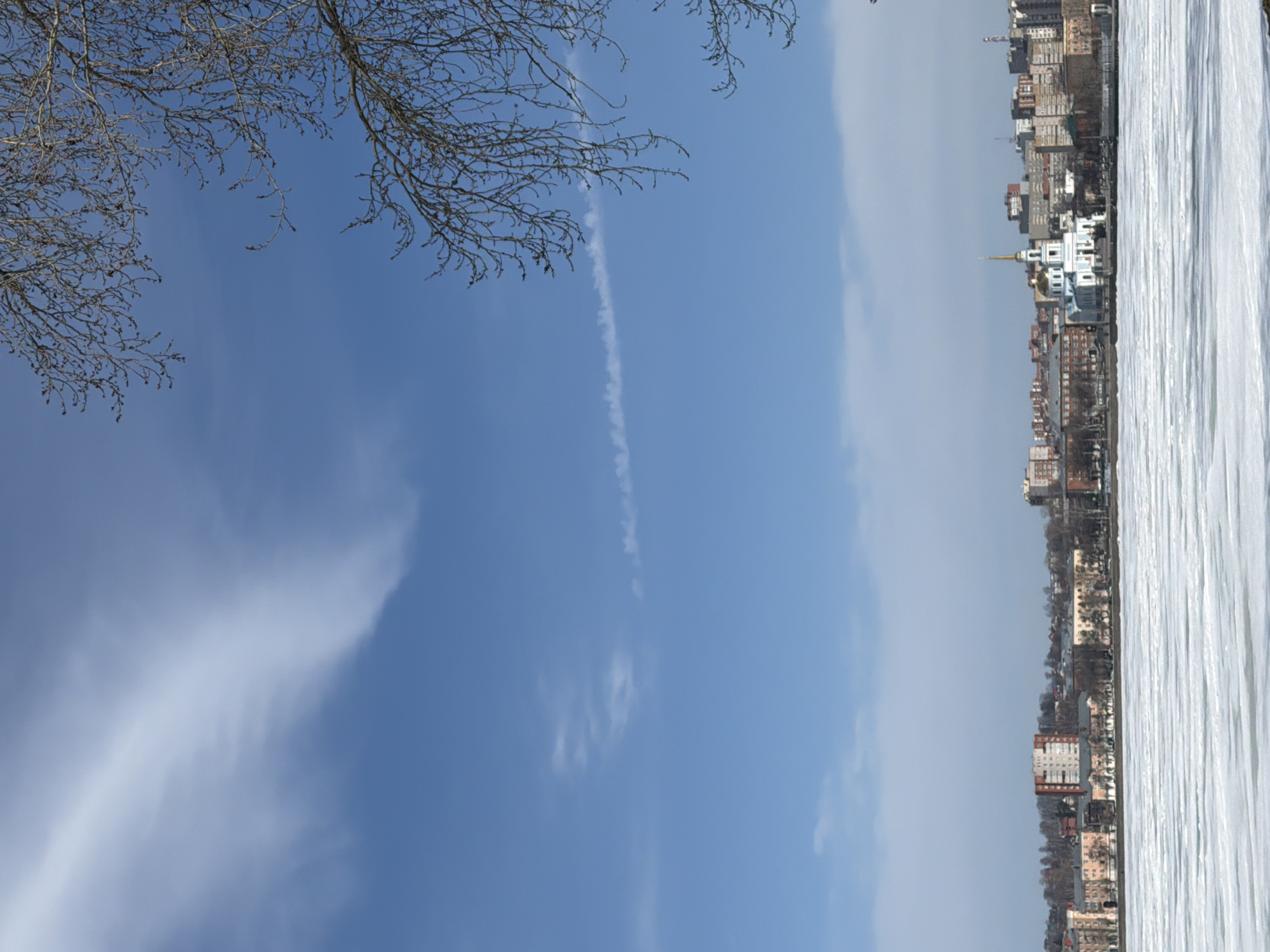 A daytime, slightly aerial shot of the city of Votkinsk, Russia. The landscape is divided into thirds horizontally.
The top left section contains tree branches against a clear blue sky, with light contrails and wispy clouds.
The middle third showcases the clear sky transitioning into a hazy, light gray area.
The bottom right section depicts a city skyline with tall buildings, dominated by a white, presumably frozen lake or body of water in the foreground. The city features a prominent church with a golden spire. The horizon line is slightly blurred due to the haze.