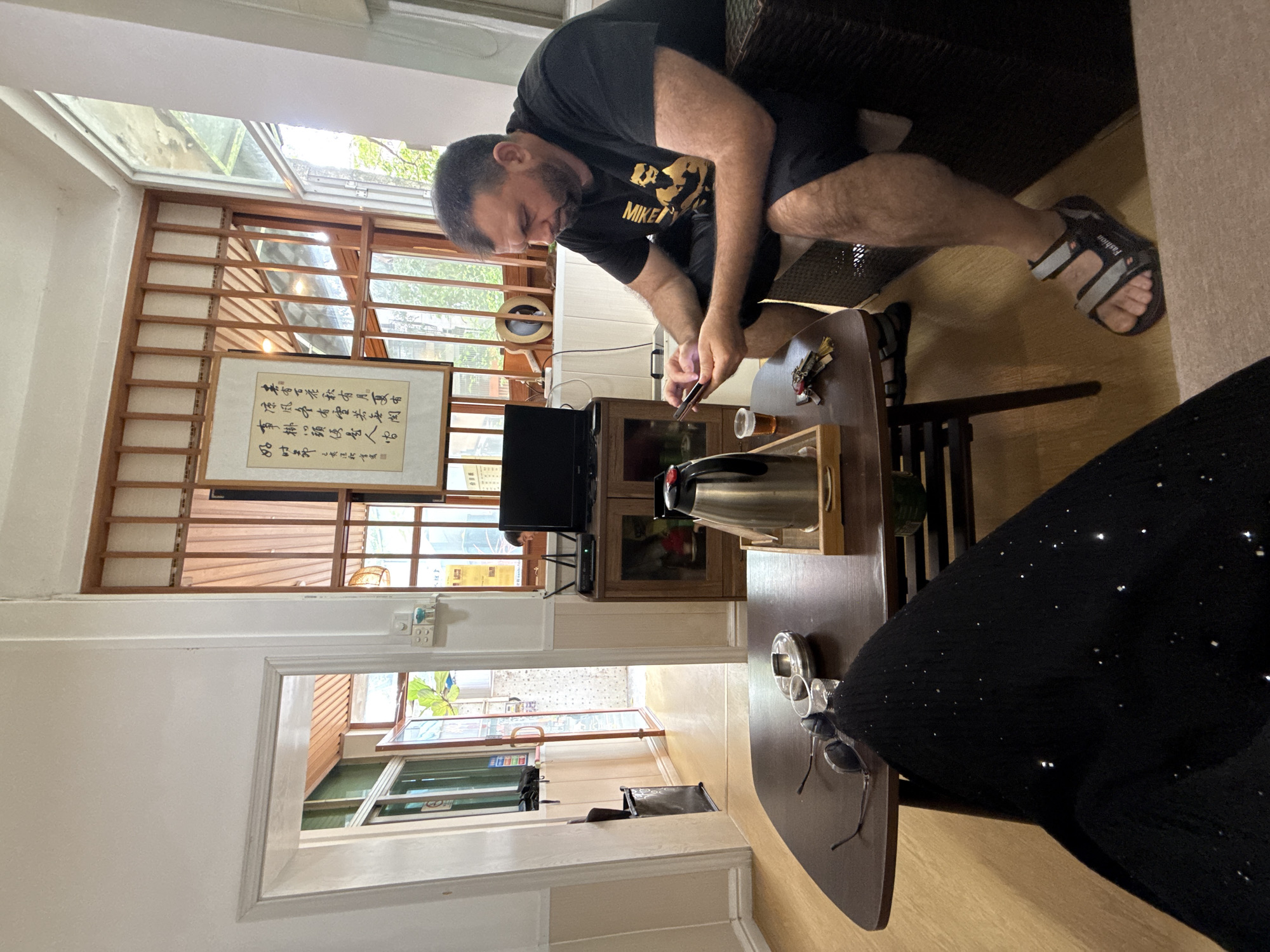 A man is sitting on a couch, looking at his phone. He is wearing a black t-shirt and black shorts. He has a beard and short hair. In front of him is a coffee table with a teapot, a glass of tea, an ashtray, and some keys. To the left of the table is a doorway leading to another room. There is a window in the background with wooden blinds. The room appears to be a living room or lounge area. The image was taken in Huizhou, China.