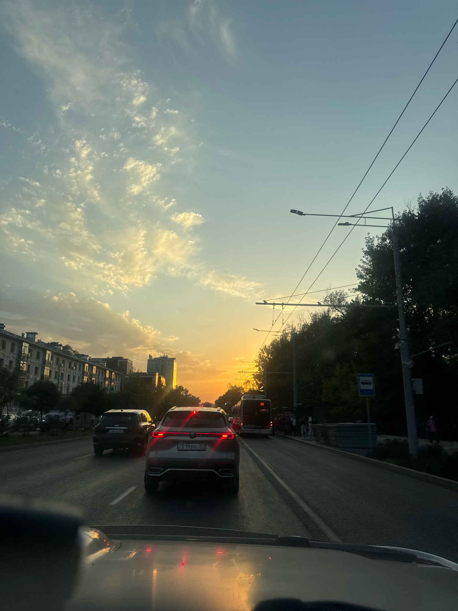 The image depicts a street scene in Almaty, Kazakhstan, during what appears to be sunset. The foreground shows the interior of a vehicle, suggesting the photo was taken from a car. The road is paved and has lane markings. Several cars are visible on the road, with one grey SUV prominently in the right lane, displaying a license plate that reads "519BVU 02". To the left, a dark-colored SUV is also on the road. Further ahead, a bus with the number "92" on its rear is stopped, and a group of people are gathered near a bus stop. The background features apartment buildings on the left, and on the right, lush green trees line the street, interspersed with streetlights and overhead power lines, typical of urban infrastructure. The sky is a mix of pale blue with scattered white and yellow clouds, illuminated by the warm hues of the setting sun, casting a golden glow on the scene. The overall impression is of a typical urban commute during the evening hour in Almaty.