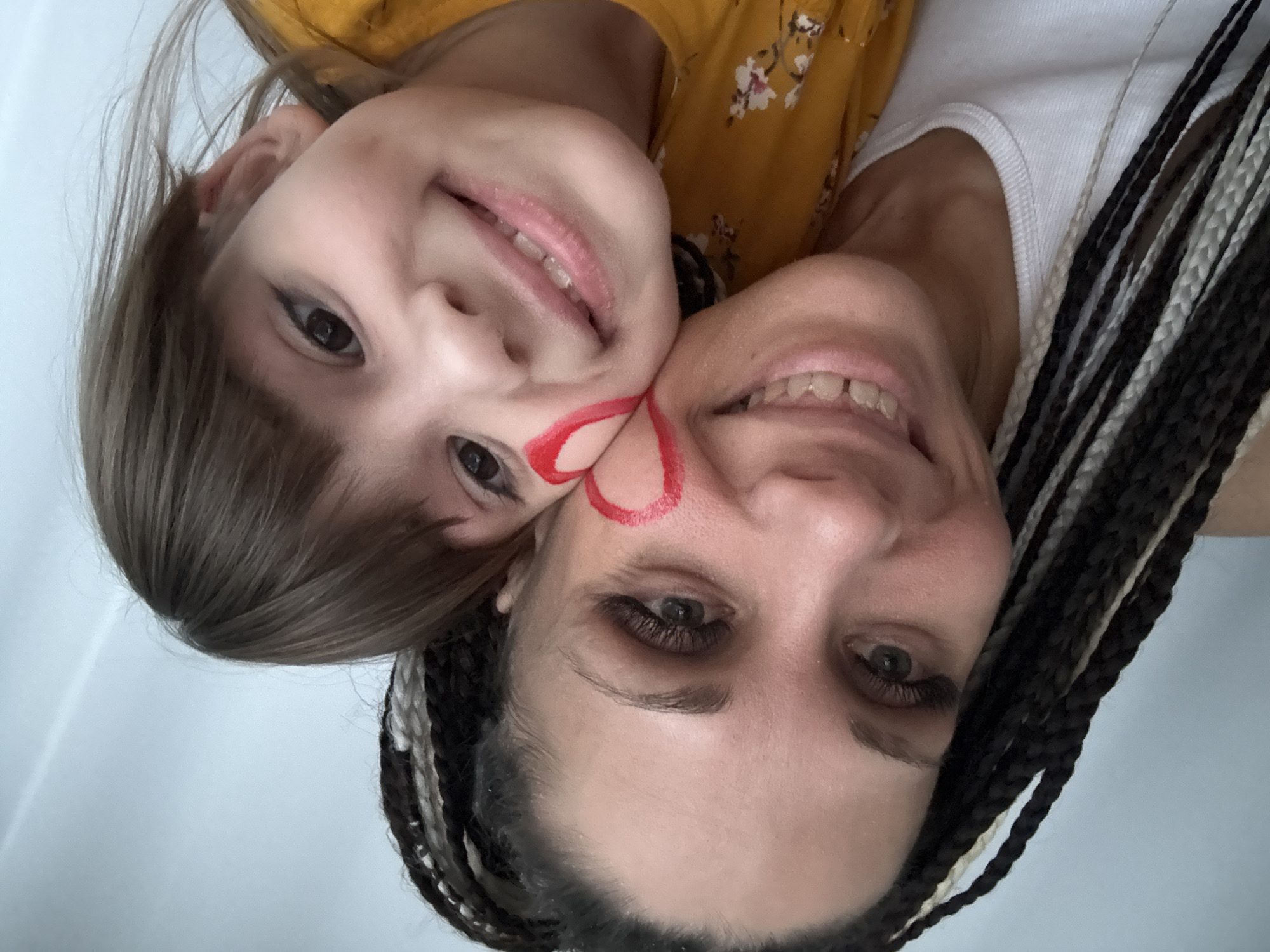 A close-up, top-down shot features an adult and a child, likely a mother and daughter, smiling broadly and positioned cheek-to-cheek. The image is rotated ninety degrees clockwise, showing their faces horizontally.
The adult, on the right, has dark hair styled in black and white braids. She wears a white tank top and has noticeable long eyelashes. Her smile reveals her teeth, and a red, heart-like shape is painted on her cheek, extending towards the child's face.
The child, on the left, has light brown hair and is wearing a yellow top with a white floral pattern. She also has a red design on her cheek that aligns with the adult's face paint, suggesting a shared artistic decoration. Both individuals are looking up towards the camera, their expressions conveying happiness and warmth.
The background is a plain, light color, possibly a wall or ceiling indoors, offering no specific location cues beyond the provided context of Rusanovo, Russia. There are no objects other than the subjects themselves, and no visible text. The interaction appears playful and affectionate, possibly a fun family moment.