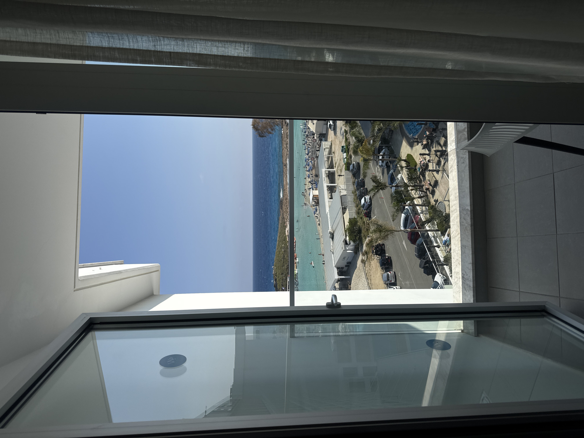 The image captures a vibrant coastal scene in Ayia Napa, Cyprus, viewed from an indoor window on a bright, sunny day. The foreground features the white frame and glass of an open window, with grey translucent curtains framing the top. A circular sticker, partially visible on the window pane, appears to show the number "21".
Beyond the window, a wide, sandy beach is visible, bustling with numerous people enjoying the clear, turquoise waters of the Mediterranean Sea. Many beachgoers are relaxing on sunbeds under a multitude of umbrellas, while others are swimming or wading in the shallow, calm sea. The water transitions from a light, clear blue near the shore to a deeper blue further out.
Adjacent to the beach runs a paved area that serves as a road or parking lot, with several cars, including SUVs and sedans, parked along its edge. Palm trees and other greenery are interspersed along this coastal strip. Modern buildings, likely hotels or beach clubs, line the area. On one prominent white building, the text "LUMA" is clearly visible, with "nissi bo" partly legible beneath it, likely referring to the famous Nissi Beach or Nissi Bay in Ayia Napa. A white balcony railing is also visible on the right side of the view.
The clear blue sky, bright sunlight, and calm sea indicate excellent weather for outdoor and beach activities. The scene conveys a sense of a popular holiday destination.