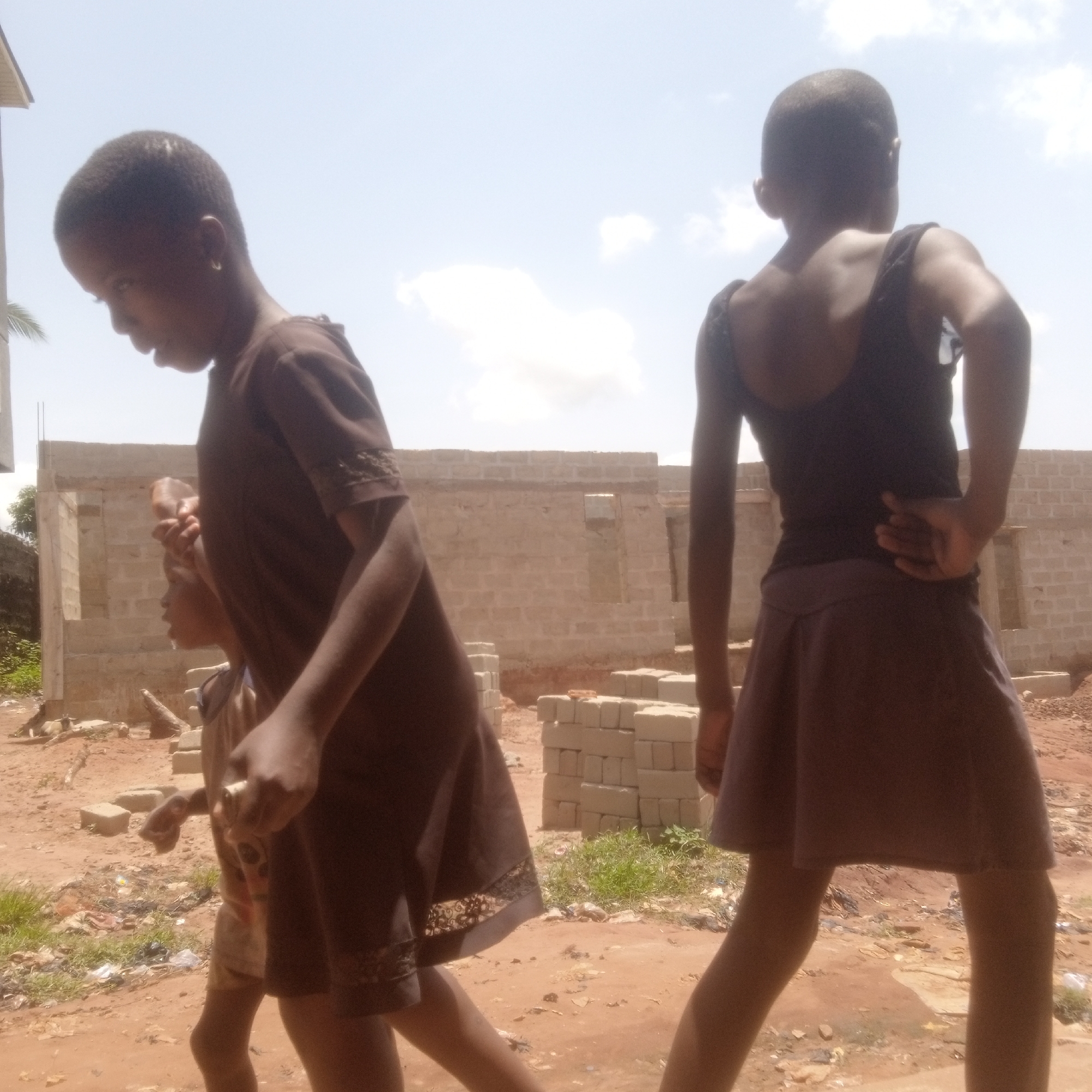 In the foreground of the image, two young girls, seen from the knees up, stand on a brown, dirt ground. One girl is facing away from the camera with her hand on her back; she is wearing a dark tank top and a brown skirt. The other girl faces to the left and is wearing a brown dress. Between them and slightly behind is a younger child, partially obscured, holding something in his hand. In the background is an unfinished brick building. The environment consists of dirt, scattered bricks, grass, and bits of trash. The location is Ukwu-Abwa, Nigeria. The sky is partly cloudy.