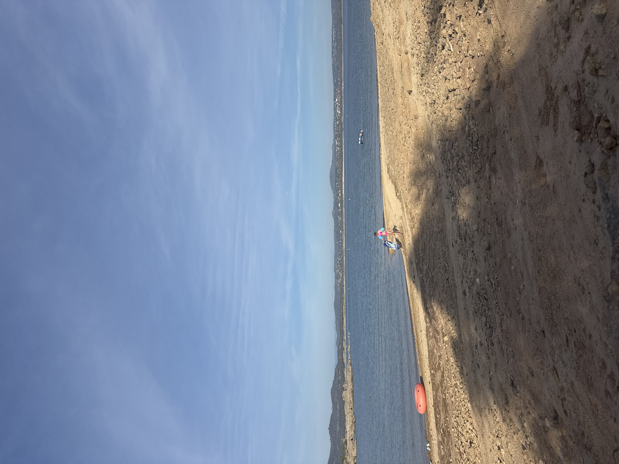 A wide shot captures a sunlit beach area in Granite Bay, California, bordering a calm lake. The foreground is dominated by dry, sandy, and rocky terrain, with shadows from unseen trees stretching across the ground. In the midground, a strip of wet sand meets the clear blue water. A bright orange inflatable ring rests on the wet sand near the water's edge. Further out in the lake, a person is visible on a raft or small boat, and on the shore, a child in pink swimwear walks away from the water, carrying a blue and white boogie board and a kickboard. The background consists of a distant shoreline with hills and scattered houses, all under a vast, pale blue sky with thin, wispy clouds.