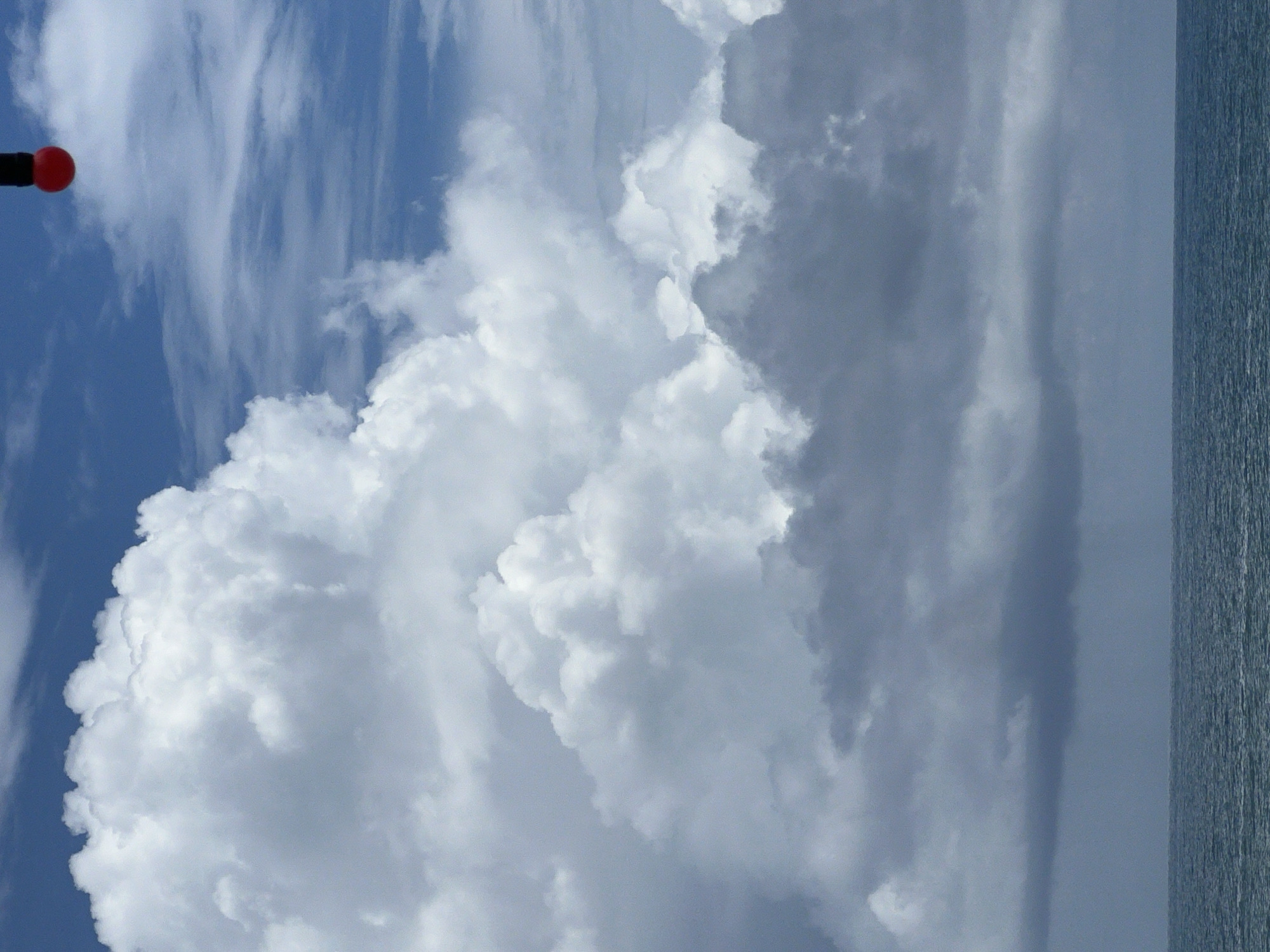 The image depicts a view of the ocean and sky. The sky is partly cloudy with a mix of white and dark gray clouds. A small red and black object is visible in the upper right corner of the frame, hanging from above. The ocean occupies the bottom portion of the image, appearing choppy and dark blue. The horizon line is clearly defined, separating the sky and the sea. The location is identified as Unknown, Unknown.