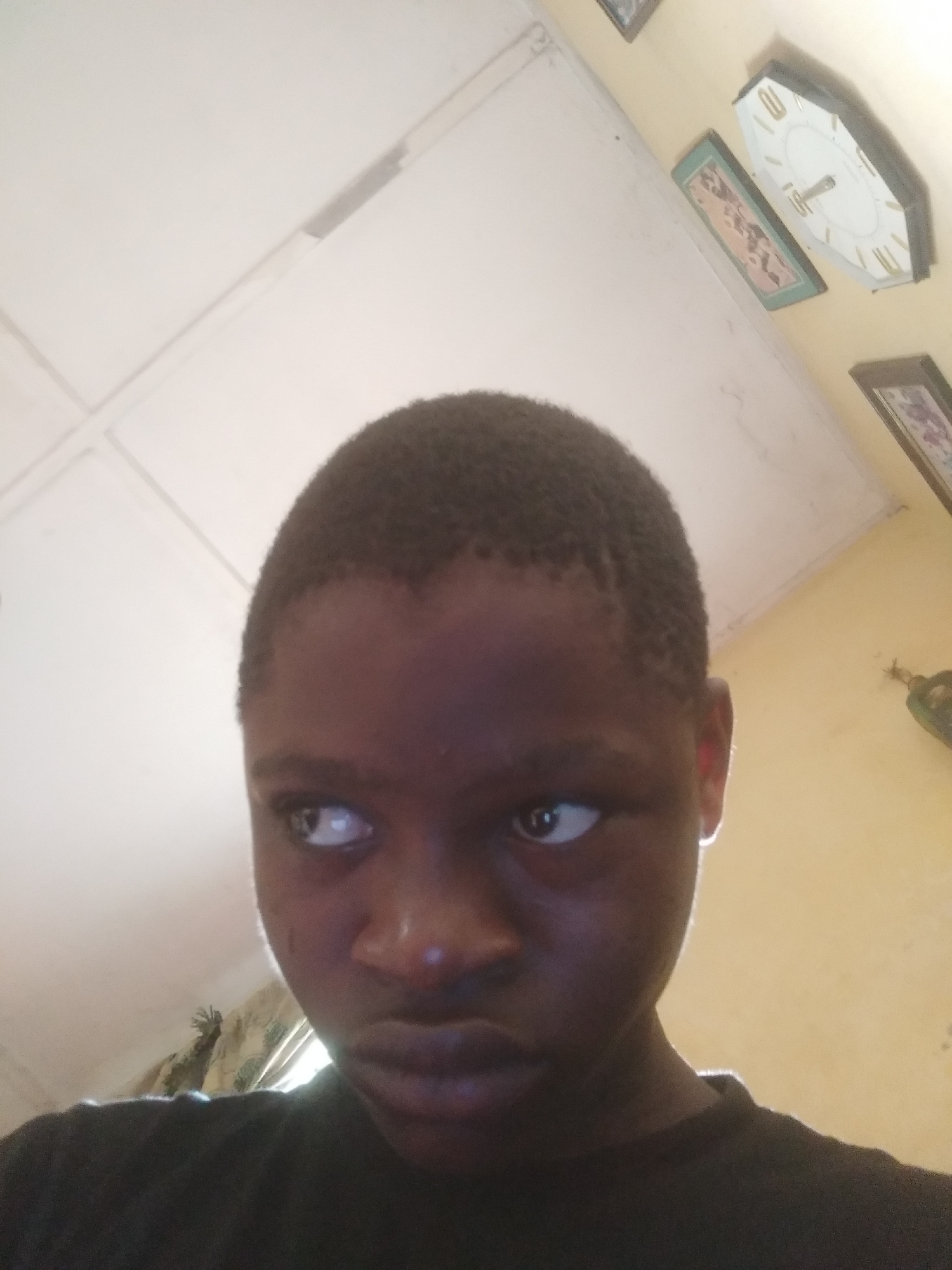 A close-up, eye-level shot captures a young person, likely male, with dark skin and short, dark hair. The individual is positioned centrally in the frame, wearing a dark, possibly black, t-shirt. Their gaze is directed sharply to the viewer's left (their right), conveying a serious, intense, or contemplative expression.
The background reveals an indoor setting. Above the person's head, a white paneled ceiling is visible, characterized by discernible seams, some of which show slight discoloration or wear. To the right of the person's head, a cream or light yellow-colored wall is adorned with several decorative items. These include an octagonal wall clock with a white face and gold-colored numerals and hands, and at least two framed pictures, one of which has a prominent green frame. A glimpse of patterned fabric, possibly curtains, can be seen in the lower left background. The lighting appears ambient and diffused, indicating an indoor environment, though no specific light source is visible. No text is clearly legible on any of the visible objects.