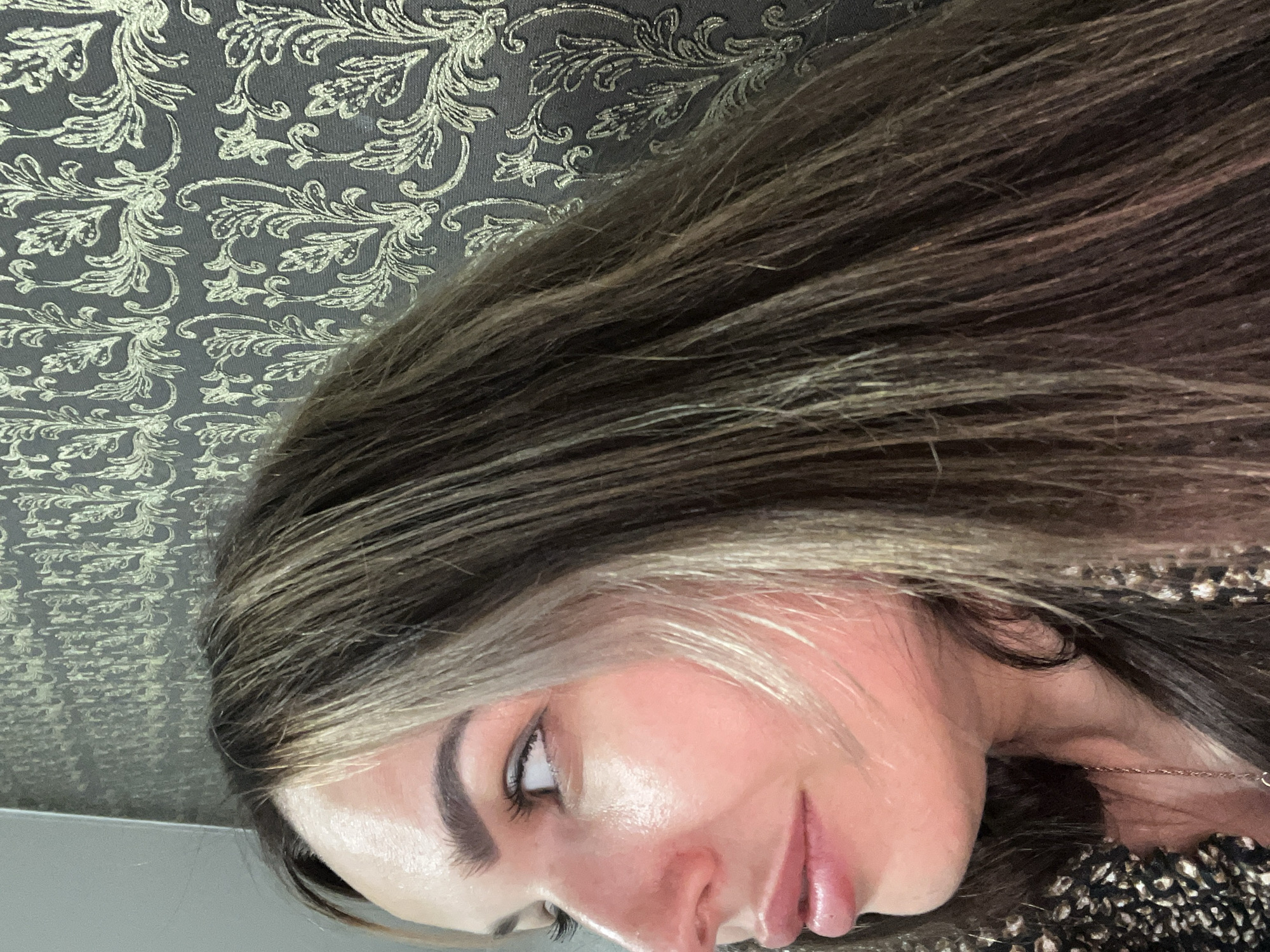 A side profile close-up shot focuses on the left side of a young woman's face. She has light skin with a warm undertone and appears to be looking to her left. Her hair, a mix of dark brown and lighter highlights, cascades down her shoulder and partially obscures the patterned wallpaper behind her. The wallpaper is a dark gray with a repeating ornate floral design in a lighter, metallic color. A portion of her patterned top and a delicate chain necklace are visible on the lower left side of the frame. The lighting highlights her cheek and the sheen on her skin.