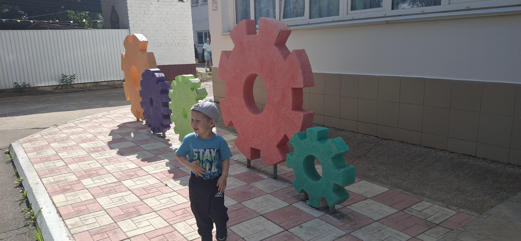 A young boy wearing a grey cap, a blue t-shirt with the word "STAY" and black sweatpants stands in front of a line of large, colorful gear sculptures. The gears are orange, purple, green, red, and teal, and are arranged in a row. The boy is smiling and looking to his left, with his right hand on his stomach. He is standing on a pathway paved with pink and beige square tiles.
Behind the boy and the gears is a building with a white brick facade and large windows. In the background, a white fence and some greenery are visible. A person in a light blue shirt and shorts can be seen walking in the distance. The scene appears to be set outdoors during the day, under sunny weather conditions. The overall atmosphere is cheerful and playful, suggesting a public space like a playground or park. The visible text on the boy's pants reads "adidas". The location is Chaikovsky, Russia.