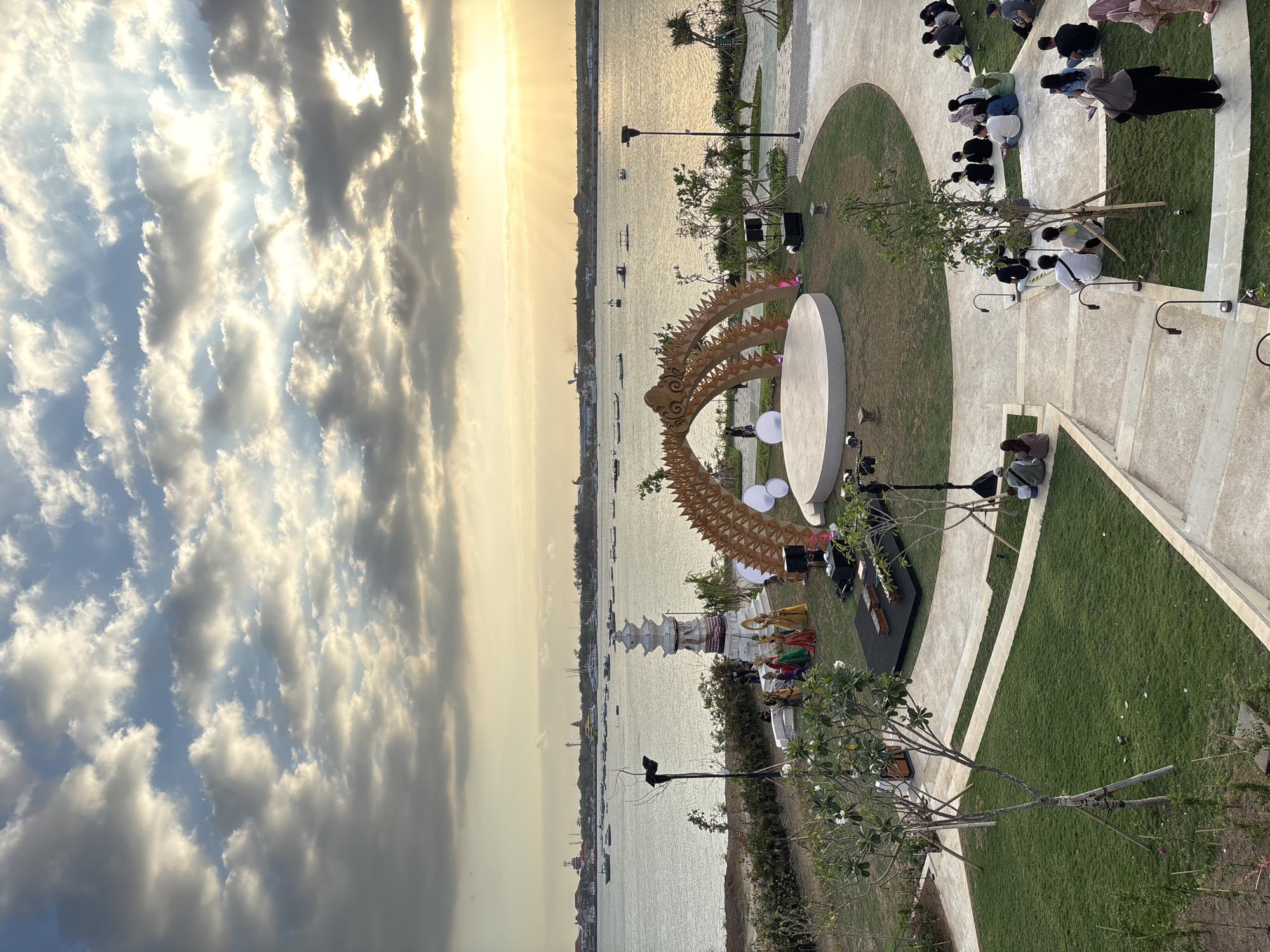 In an outdoor amphitheater setting overlooking a body of water, a sunset casts a warm glow through dramatic clouds. The foreground features a circular stage adorned with a golden, arching, decorative structure and several large white orbs. To the left of the stage, a group of individuals in colorful traditional attire are gathered near a multi-tiered white monument. The amphitheater is equipped with lighting fixtures and sound equipment. Spectators are seated on the grass and tiered concrete steps, facing the stage. In the background, boats dot the water, and a distant cityscape is visible. The overall scene suggests an outdoor performance or cultural event in progress.
