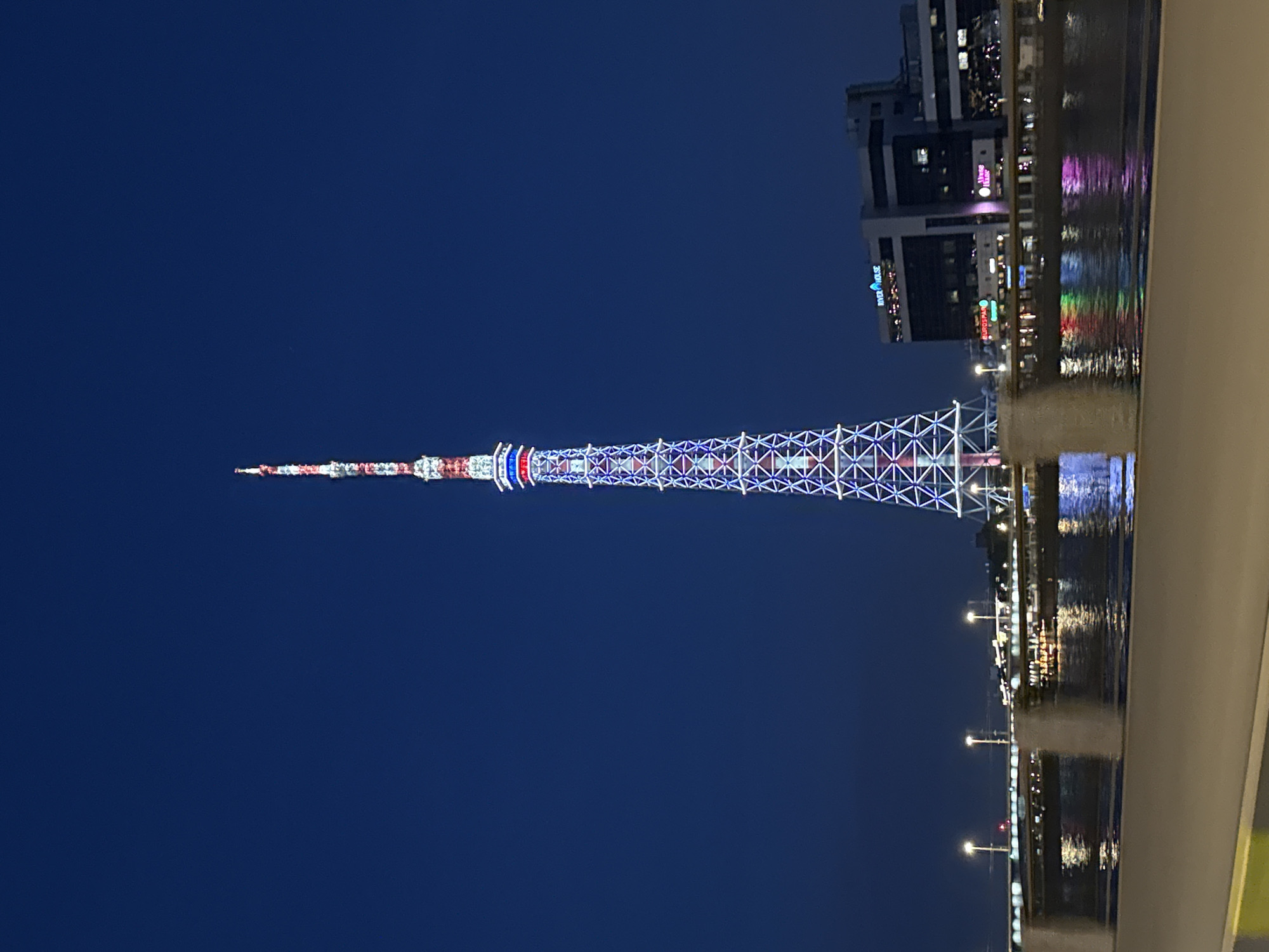 This night-time photograph, taken in Saint Petersburg, Russia, showcases the strikingly illuminated Saint Petersburg TV Tower against a dark blue sky. The scene features the towering structure, a body of water, and buildings along the opposite bank, all adorned with artificial light.
The central subject is the tall, slender television tower, characterized by its intricate lattice-work design. It is brightly illuminated in sections with red, white, and blue lights, creating distinct patterns along its height; the lower and middle sections display a grid of blue and white lights, while the upper part features red and white stripes leading to a pointed top.
In the foreground, a calm body of water, likely a river or canal, reflects the colorful lights from the tower, the streetlights, and the buildings, creating shimmering streaks of light on its surface. Along the far bank, several modern, multi-story buildings are visible. Many of their windows are lit, and some display illuminated signs. Notably, "RIVER HOUSE" is visible in blue neon, and "BURGER LAB" in red text can be seen on one building. Another partially visible sign appears to say "WINGS". A series of streetlights casts warm glows along the water's edge.
There are no people or specific activities depicted in the image. The clear night sky provides a stark backdrop for the vibrant urban illumination. The overall impression is one of a peaceful yet dynamic cityscape at night.