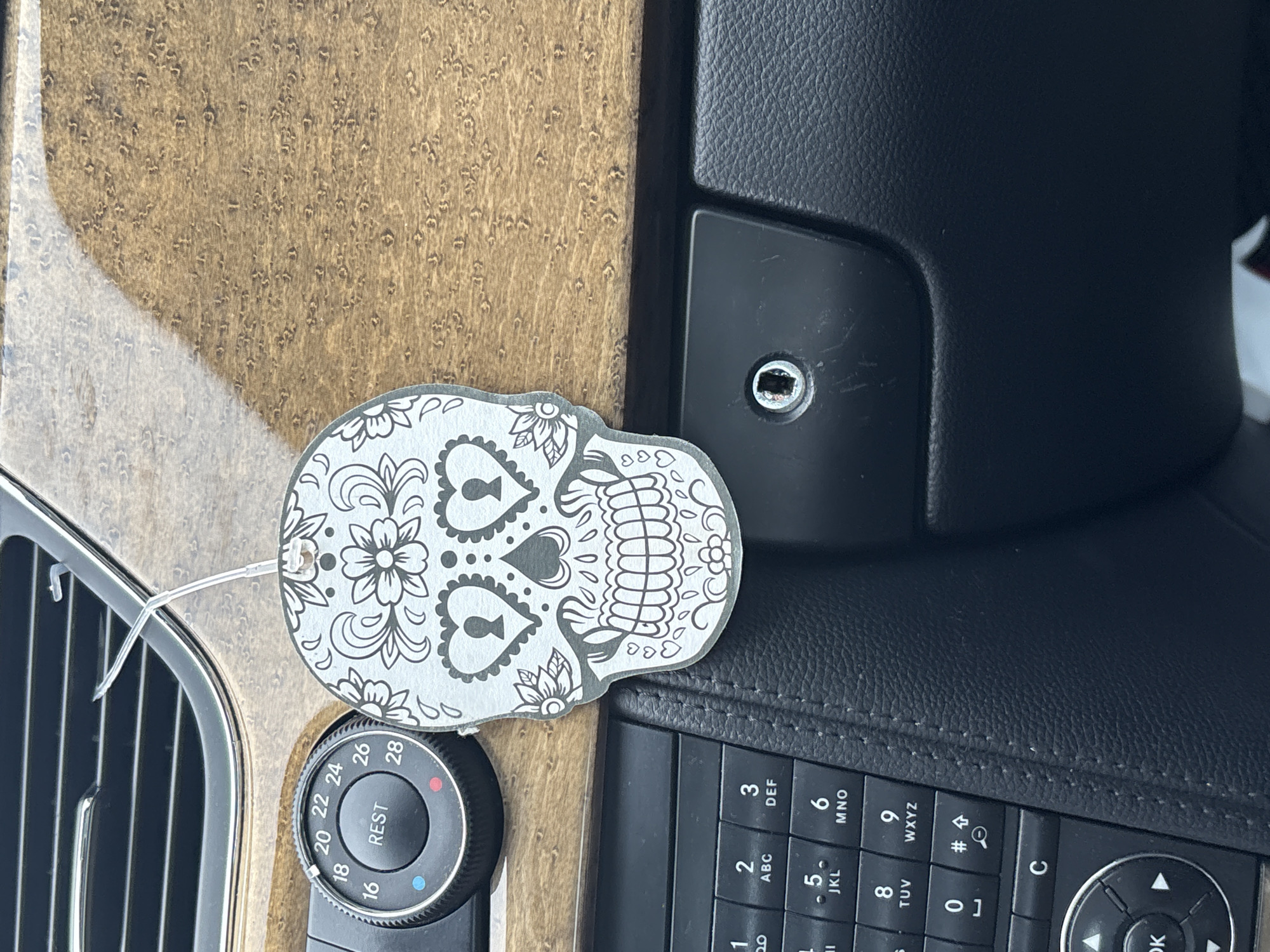 The image shows a close-up view of a car's interior dashboard. In the center, a skull-shaped air freshener with decorative floral patterns is hanging from an air vent. To the left of the air freshener is a circular dial with temperature settings ranging from 16 to 28, labeled "REST" in the center. Below the air freshener and dial is a section of the car's center console, featuring a black, textured surface and a keypad with numbers and symbols. The background is dominated by the wood-grain texture of the dashboard. The scene is well-lit, likely by daylight. There are no people visible in the image. This scene is not in Гацукоўскі сельскі Савет, Belarus, as it depicts a car interior.