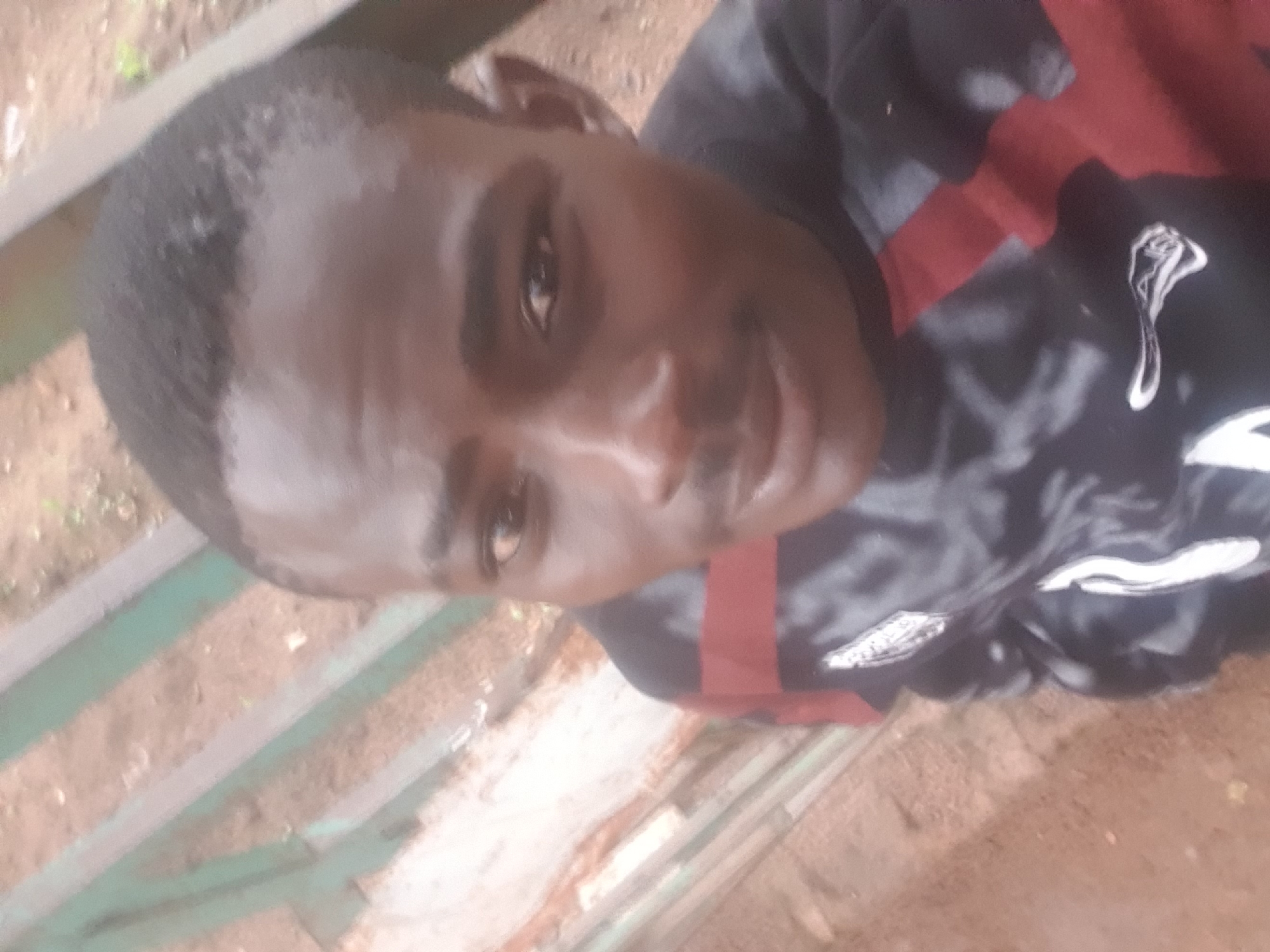 A close-up, outdoor, selfie-style photograph shows a young Black male looking at the camera. He has dark skin, short dark hair, dark eyes, and a faint mustache. He is wearing a black and red patterned long-sleeved shirt with white accents. The background appears to be a park or outdoor area with a dirt path and a faded green and brown wooden fence. The lighting is soft and natural. The image is slightly blurry, indicating movement during the capture. The specific location in Washington, United States of America, cannot be determined from the visual information provided.