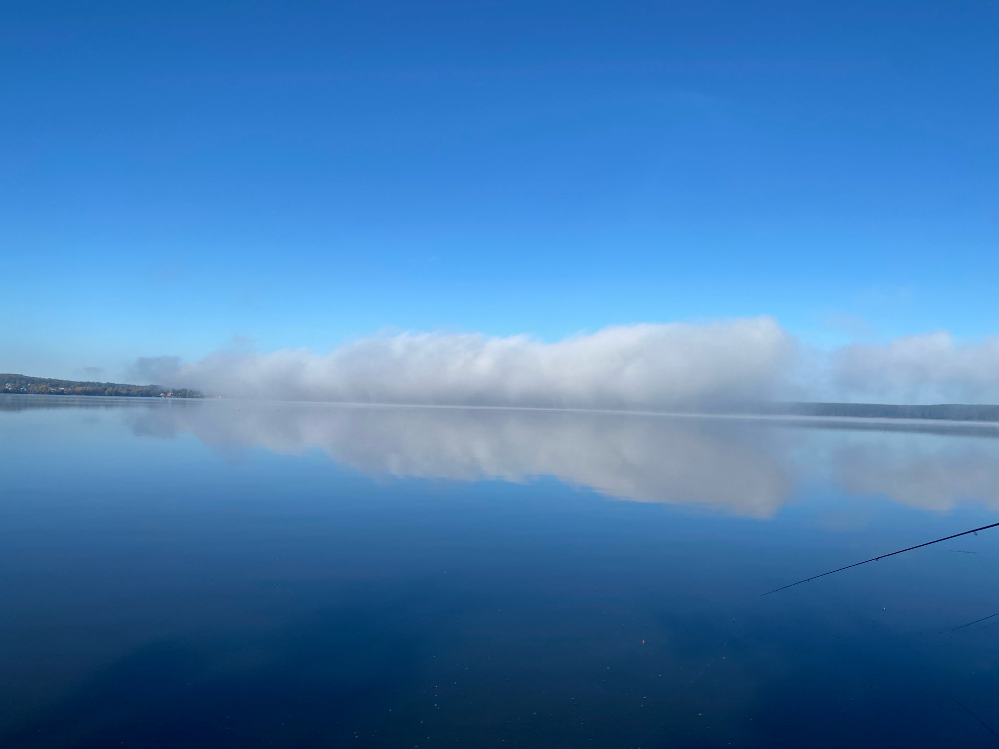 The image depicts a tranquil scene on a calm lake, likely in or near Votkinsk, Russia, on a clear, bright day. The sky is a vast expanse of uninterrupted blue, with a thin, horizontal band of white fog or low-lying clouds stretching across the middle distance, partially obscuring the distant shoreline. This fog is reflected with striking clarity on the glassy surface of the water, creating a mirrored effect.
The water itself is a deep, rich blue, reflecting the sky and fog. Scattered across its surface are small, subtle disturbances, possibly ripples from the gentle breeze or the movement of aquatic life. In the foreground, on the right side of the frame, a fishing rod is visible, its tip dipping slightly towards the water. The rod suggests the presence of a person, though no individuals are directly visible in the frame. A small, orange float is also discernible on the water's surface near the fishing rod.
The distant shoreline is low-lying and appears to be lined with trees, with some faint indications of buildings or structures nestled amongst them, consistent with a lakeside settlement. The overall impression is one of serene, natural beauty, with a hint of human activity represented by the fishing rod.