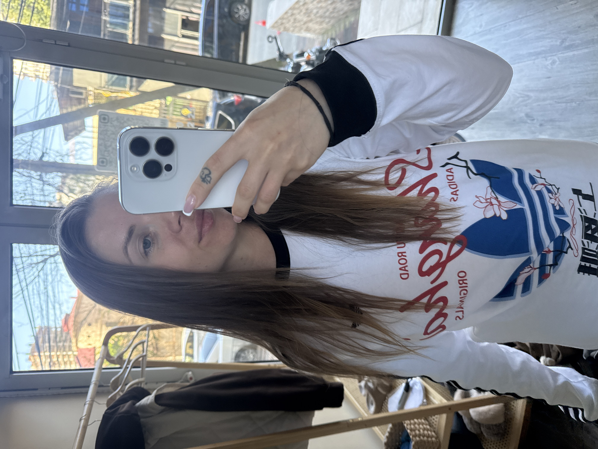 A young woman with long brown hair is taking a mirror selfie indoors, in what appears to be a room or hallway in Batumi, Georgia. The image shows her from the chest up, holding a white iPhone with three cameras in front of her face, partially obscuring it. She is wearing a white long-sleeved shirt with black cuffs, which features a prominent graphic design and text.
The reflections in the large mirror reveal an outdoor urban scene with buildings, trees, and what appears to be a street with a car and a motorcycle. The sunlight suggests it is daytime. A clothing rack with various garments is visible behind her in the mirror's reflection. Her expression is neutral, and she has natural makeup. A small, indistinct tattoo is visible on her right index finger, and her nails are manicured with French tips.
Visible text on her shirt, which is mirrored due to the reflection, includes "ADIDAS," "ORIGINALS," and "ROAD," along with other stylized red and blue graphics. A QR code is also faintly visible in the reflection of the window.