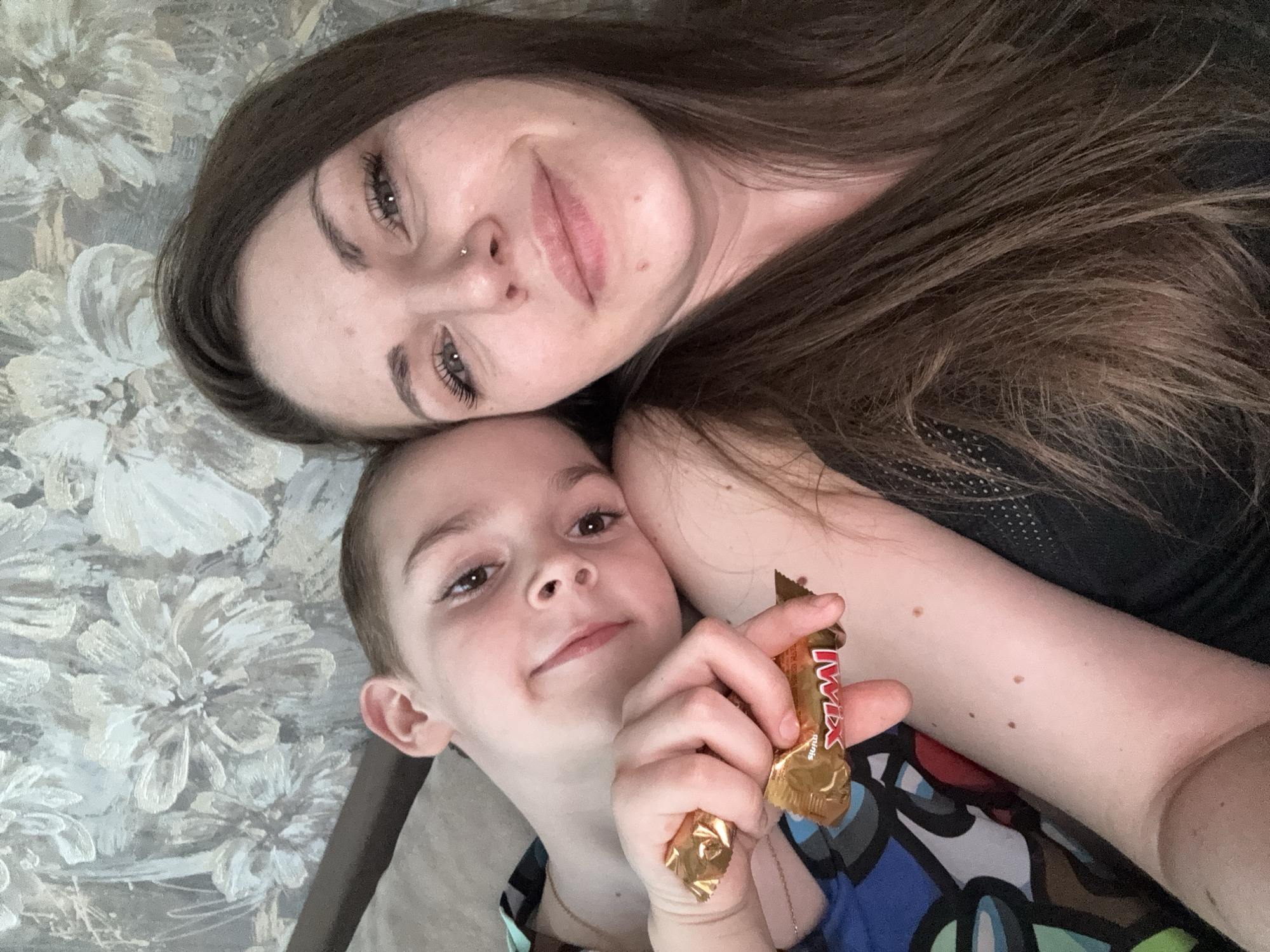 A woman and a young boy are lying down next to each other in a selfie taken indoors. The woman is at the top of the frame with her head resting on the boy's. She has long, dark brown hair, fair skin, and wears a nose piercing. The boy is below her, holding a partially unwrapped Twix chocolate bar in his hand. He looks directly at the camera with a slight smile. The background features a floral pattern. The location is Раздолье, Russia.
