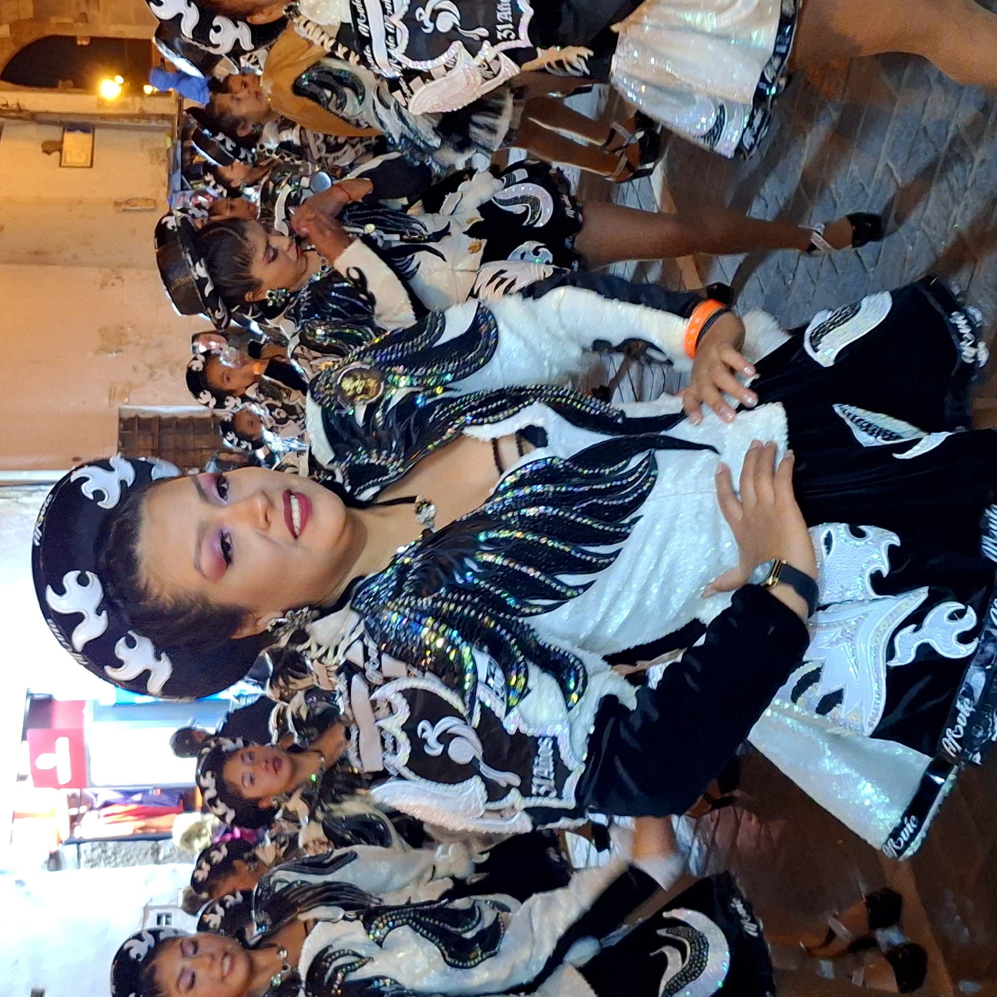 The image shows a group of women in elaborate, traditional costumes, possibly during a parade or cultural event, in what appears to be an urban setting at night. The central figure, a woman with dark hair and striking makeup, is the main focus. She wears a black and white patterned hat adorned with white abstract designs, a black and white sequined jacket with dramatic, wing-like shoulder pieces, and a white top with black lacing. Her costume features intricate embroidery and metallic accents. Other women in similar costumes are visible in the background, some with hats and decorative jackets. The ground appears to be paved with cobblestones. The environment suggests an outdoor street scene, with blurred buildings and lights in the background. The overall impression is one of a vibrant cultural celebration.
