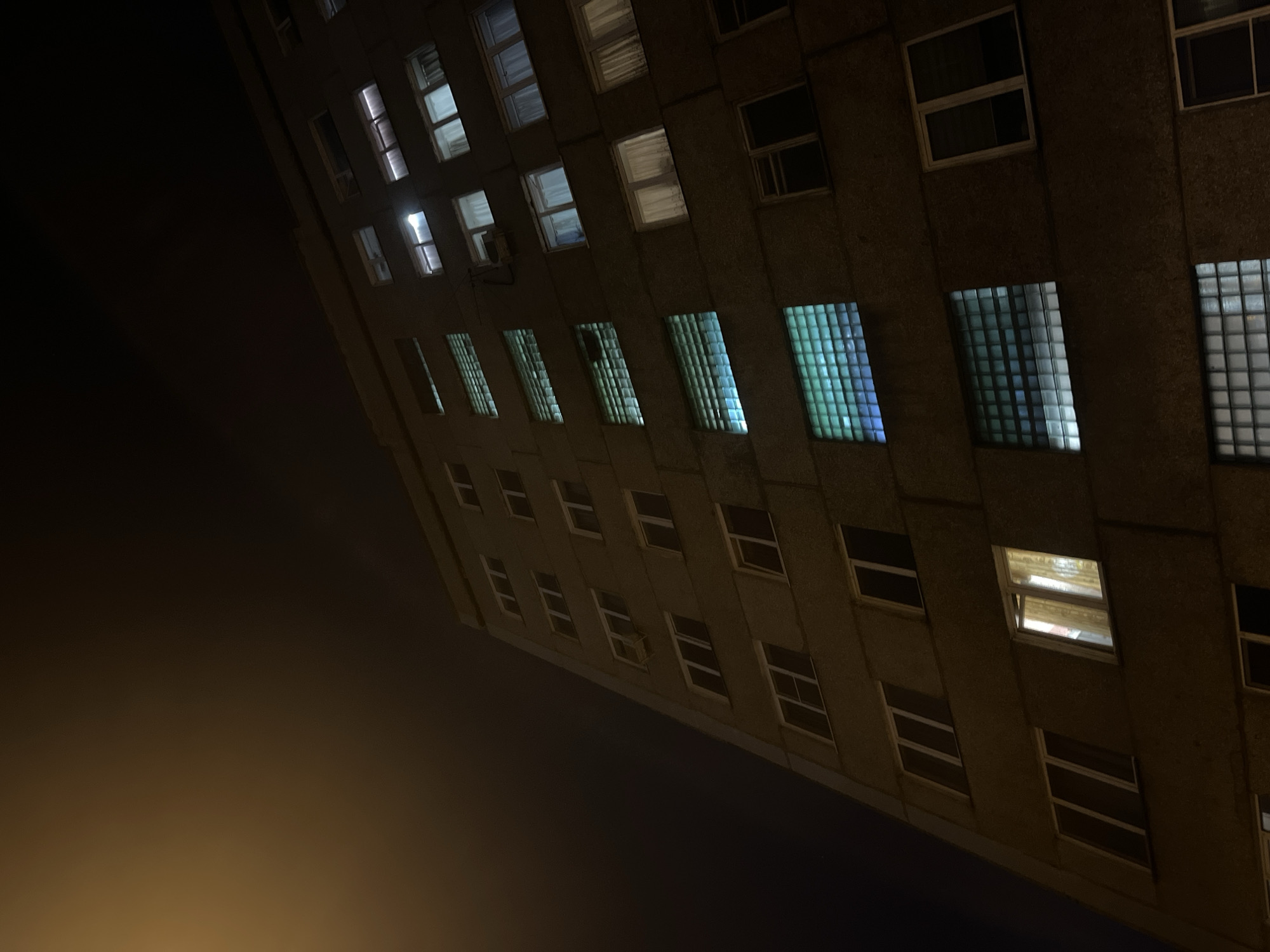 A low-angle view captures a building at night, possibly located in Krasnoyarsk, Russia, as inferred from the architecture and urban setting. The structure is illuminated by lights emanating from several windows, creating a contrast against the dark sky. The building's exterior appears to be concrete or a similar material. The foreground is dark, suggesting a nighttime environment, with a subtle gradient of light at the bottom, possibly indicating a street light or other nearby source.
