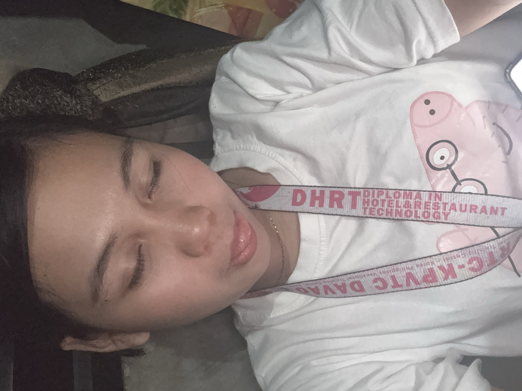 The image is a close-up, head-and-shoulders shot of a young woman with her eyes closed and her tongue sticking out slightly. She is wearing a white t-shirt with a printed image of Peppa Pig on it. A lanyard is around her neck, with text that appears to read "KPVTC DAVAO" and "DHRT DIPLOMA IN HOTEL & RESTAURANT TECHNOLOGY." The background is dark and indistinct, suggesting an indoor setting. The text on the lanyard indicates the image was taken in Davao City, Philippines, specifically referencing the Davao Region, Korea Philippines Vocational Training Center (KPVTC) and the Davao Hotel and Restaurant Technology (DHRT).