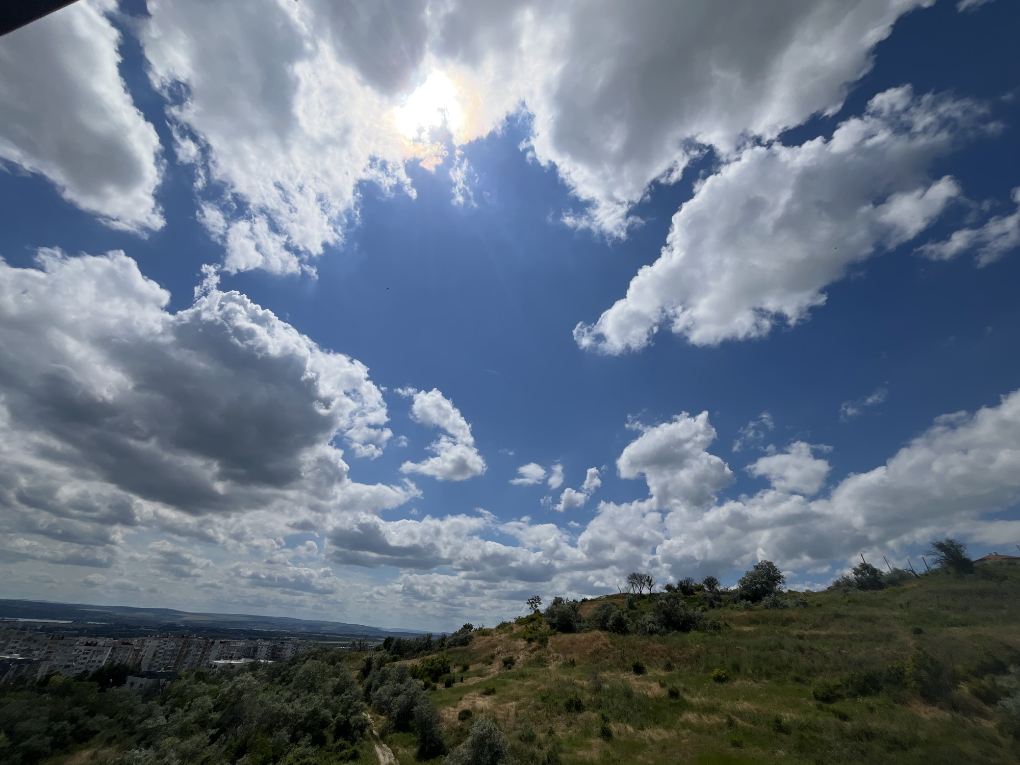 The image captures a panoramic view from a hillside overlooking the city of Varna, Bulgaria. The sky is a vibrant blue, dotted with fluffy white cumulus clouds. The sun is partially obscured by a cloud formation in the upper center of the image, casting a bright glow and lens flare. In the foreground, a grassy, undulating hillside is covered in scattered trees and shrubs. A dirt path winds its way down the slope. In the middle ground, the cityscape of Varna is visible, characterized by rows of apartment buildings. The background shows a hazy landscape with distant hills. The overall impression is one of a bright, sunny day with a mix of natural and urban scenery.