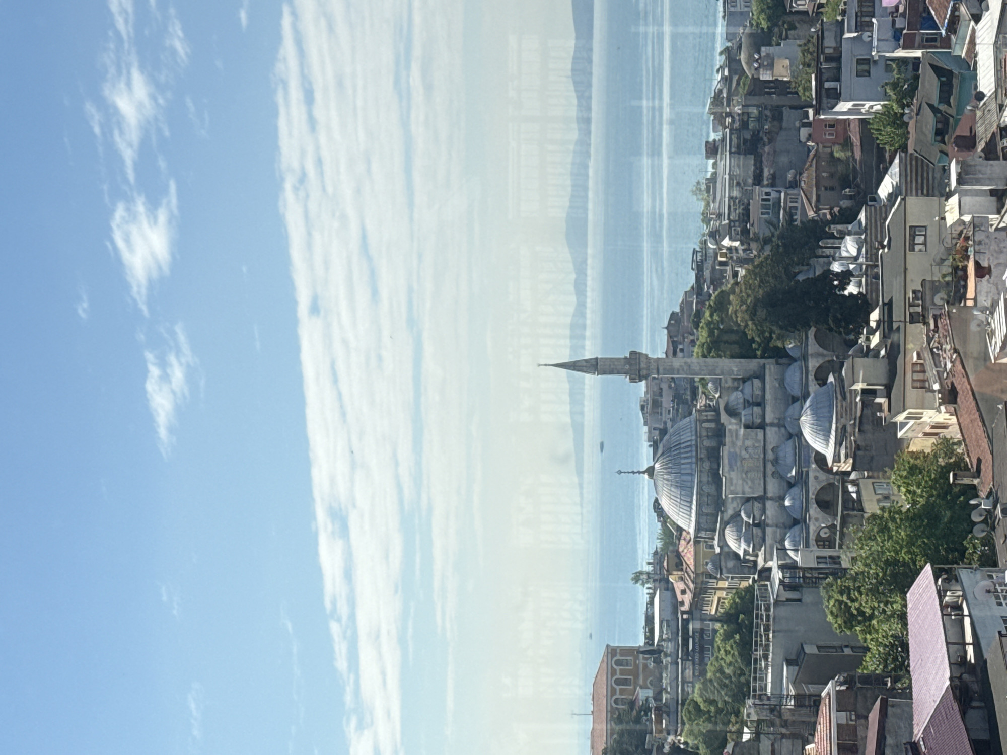 Here is a detailed description of the image:
The image presents a view of a cityscape with a large body of water in the background, suggesting a location by a sea or a large lake. The sky is visible on the left side of the frame, marked with a mix of blue and cloud cover.
In the foreground to midground, a dense urban setting can be observed. There is a prominent mosque or a similar religious building with domes and a minaret. Adjacent to the religious structure, the cityscape consists of diverse buildings, ranging in height and architectural styles. These buildings are interspersed with trees, creating a mix of urban and natural elements.
The body of water in the background appears to be calm, with subtle reflections. There are hints of landforms on the distant horizon across the water. The entire scene is captured from an elevated position, possibly from a high building, allowing for a panoramic view of the cityscape and the body of water. The image overall showcases an urban environment blended with natural elements and a scenic water view.