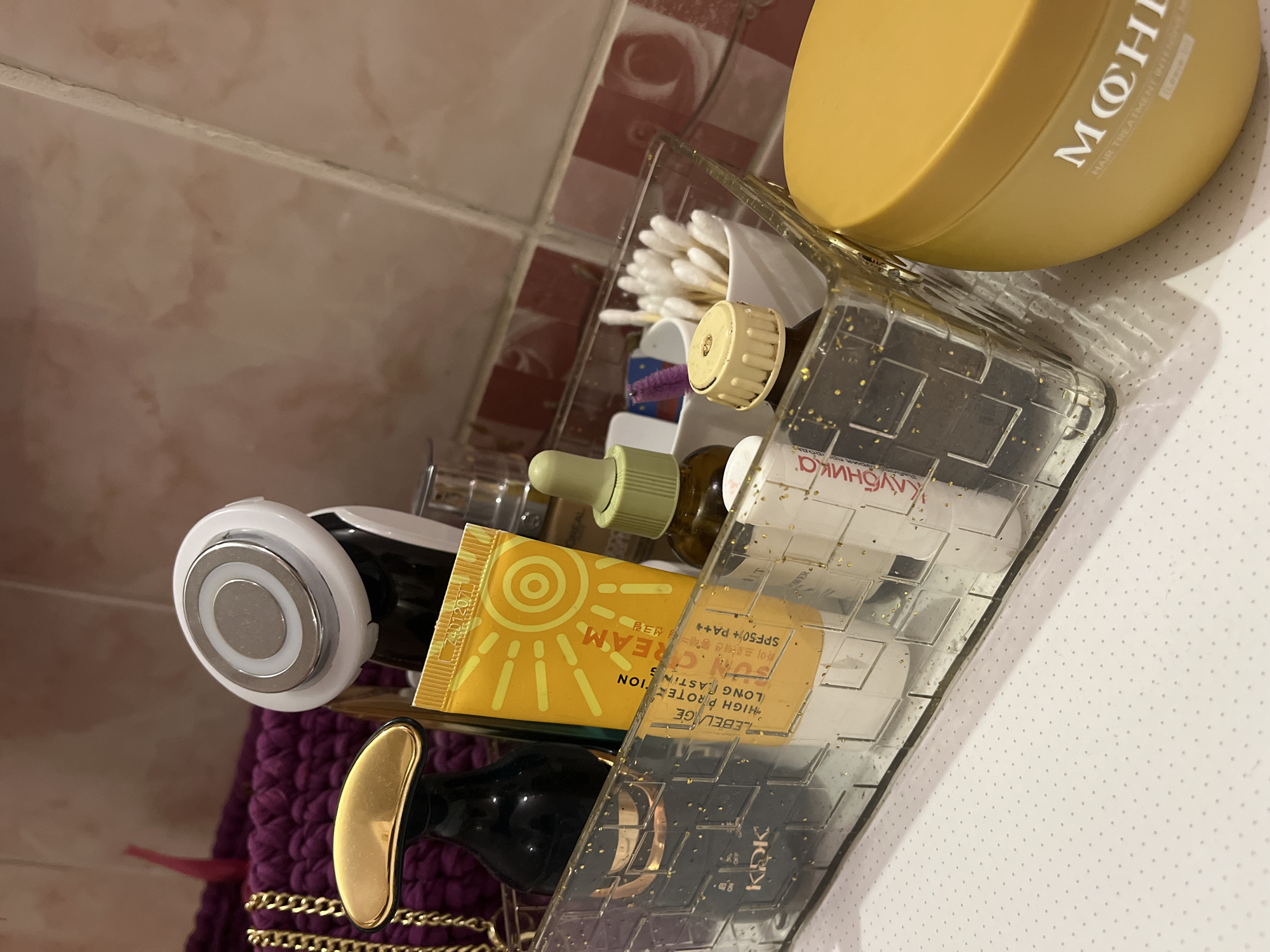 A high-angle, close-up shot depicts a clear plastic organizer filled with various personal care items. The organizer, speckled with gold flakes, contains a container of cotton swabs, a brown bottle with a dropper, a white and black facial device, and multiple tubes of creams or lotions. One visible tube is yellow with a sun icon and the words "SUN CREAM," "HIGH PROTECTION," and "LONG LASTING." The packaging also indicates "SPF50+ PA++" and the brand name "LEBELAGE." Another label reads "Клубника." A round, yellow container labeled "MOH" is positioned atop the organizer. The background includes a tiled wall with red and white checkered tiles, and a white surface with small gray dots. The scene likely takes place in a bathroom or similar area where personal care items are stored. The exact location in the city of Chaikovsky, Russia, cannot be determined from the visual information.