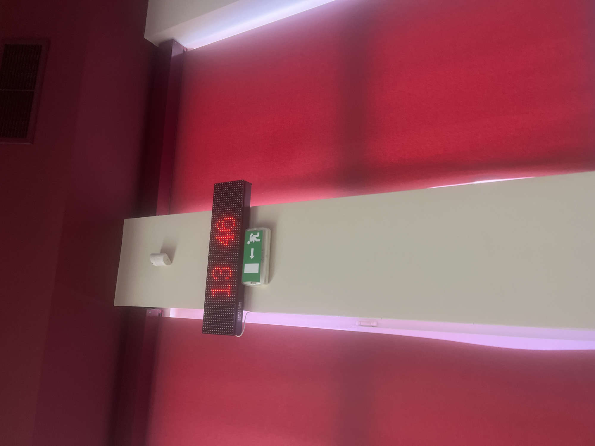 The image captures an indoor scene, dominated by vibrant red elements and a light-colored horizontal panel. A digital LED display, oriented vertically, is mounted on the left side of this light panel, prominently showing the time "19:46" in bright red digits. Text visible on the display's casing includes "LED 100 80" and a partial serial number "34-11-71".
Immediately to the right of the LED display, a green emergency exit sign is affixed to the same horizontal panel. This sign features a standard white pictogram of a running person and an arrow pointing downwards. A small, white, cylindrical object is also mounted on the left end of the horizontal panel.
The background consists of deep red blinds or curtains, which appear to be illuminated from behind, creating a soft glow along their edges. On the far left, a darker red wall features a rectangular vent or grille. The ceiling or upper wall area is light cream or white. The time displayed, 19:46, indicates it is evening. This interior scene is located in Plovdiv, Bulgaria.