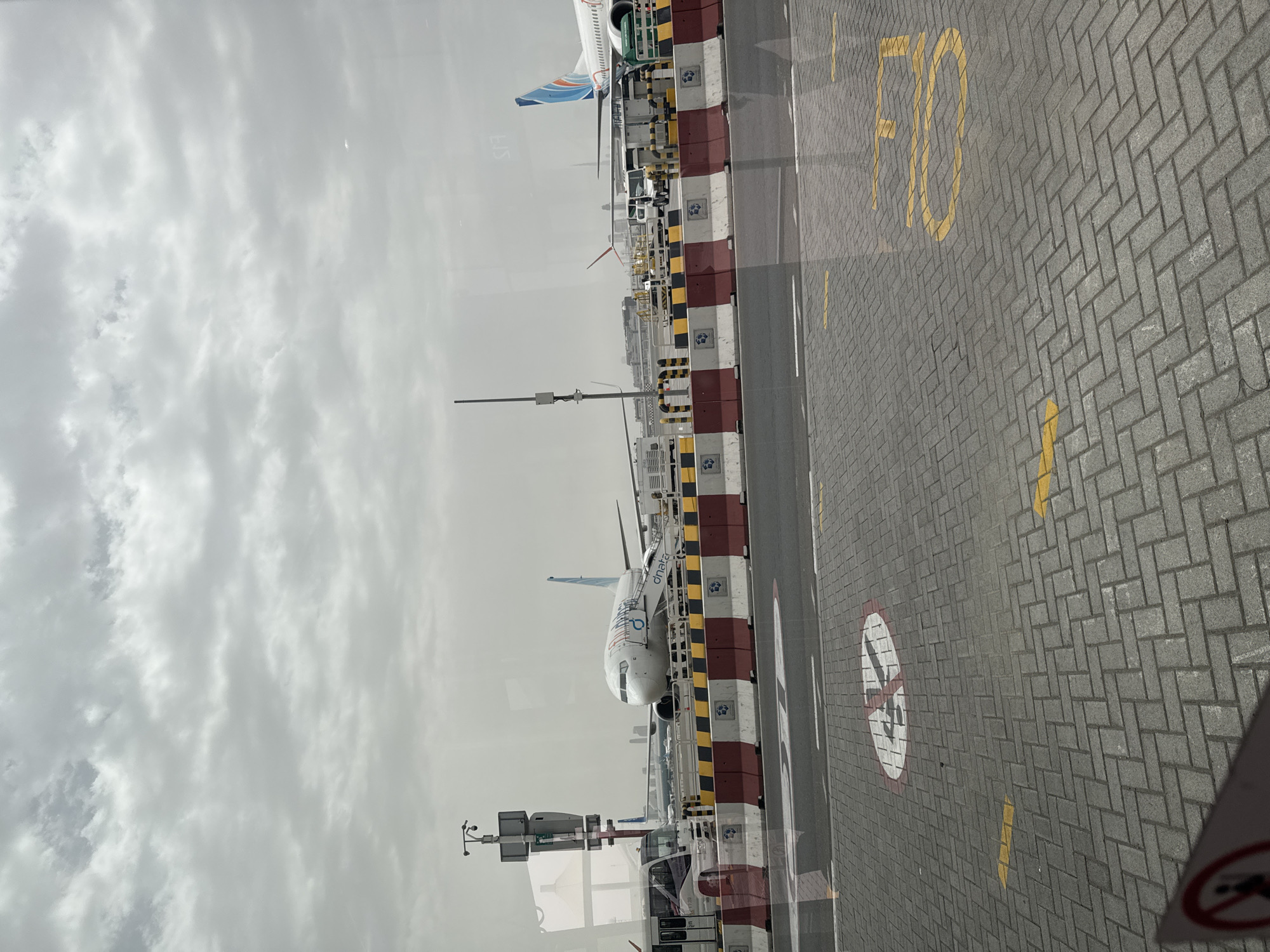 The image depicts an airport scene, likely within Dubai International Airport (DXB) in the United Arab Emirates, viewed through a window. Two aircraft are visible on the tarmac. One is closer to the camera, painted white with blue and orange details on the tail section. The other aircraft is further away and only the tail is visible. Ground vehicles and equipment are parked near the aircraft. The ground is paved with interlocking grey bricks and is marked with yellow lines and painted designations, including the number "50" and a "no smoking" symbol inside a circle. The sky overhead is overcast with dense, grey clouds.