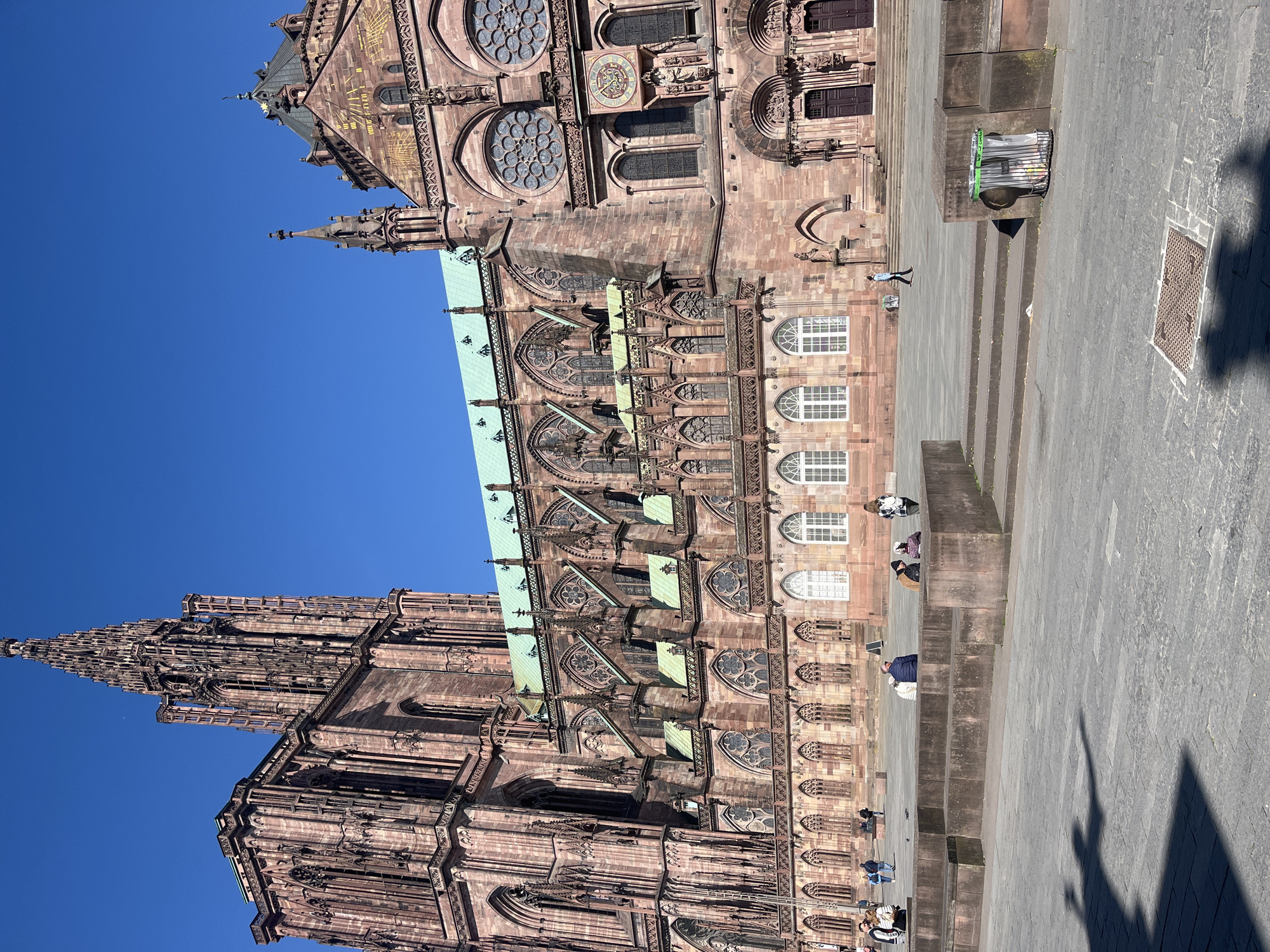 The image displays the magnificent Cathédrale Notre-Dame de Strasbourg in France, bathed in bright sunlight under a clear, vibrant blue sky. The cathedral, constructed from reddish-brown sandstone, exhibits intricate Gothic architecture with numerous detailed carvings, pointed arches, and decorative elements.
The photograph captures a broad view of the cathedral's facade, including its towering, ornate spire on the left side, which rises majestically into the sky. On the right side of the image, a different section of the cathedral, possibly part of the south transept or nave, is visible. This section features several large rose windows and a prominent astrological clock with Roman numerals. Above this, golden markings, possibly a sundial or decorative inscriptions, adorn the stone. Sections of the roof show a distinctive green patina, likely from oxidized copper.
Below the cathedral, a wide, paved public square or plaza extends. Several broad steps lead up to the building. Numerous people are scattered across this area; some are walking, while others are seated on the steps, appearing to relax or observe the surroundings. They are dressed in casual clothing.
The strong, crisp shadows cast on the light gray pavement by the cathedral and the individuals suggest a time of day with high sun, likely late morning or early afternoon. Visible objects on the ground include a black wire mesh trash bin with a green rim near the steps and a metal manhole cover. The scene conveys a sense of peaceful activity around a grand historical landmark on a beautiful, sunny day. No explicit readable text, other than the general form of the clock's numerals, is clearly visible.