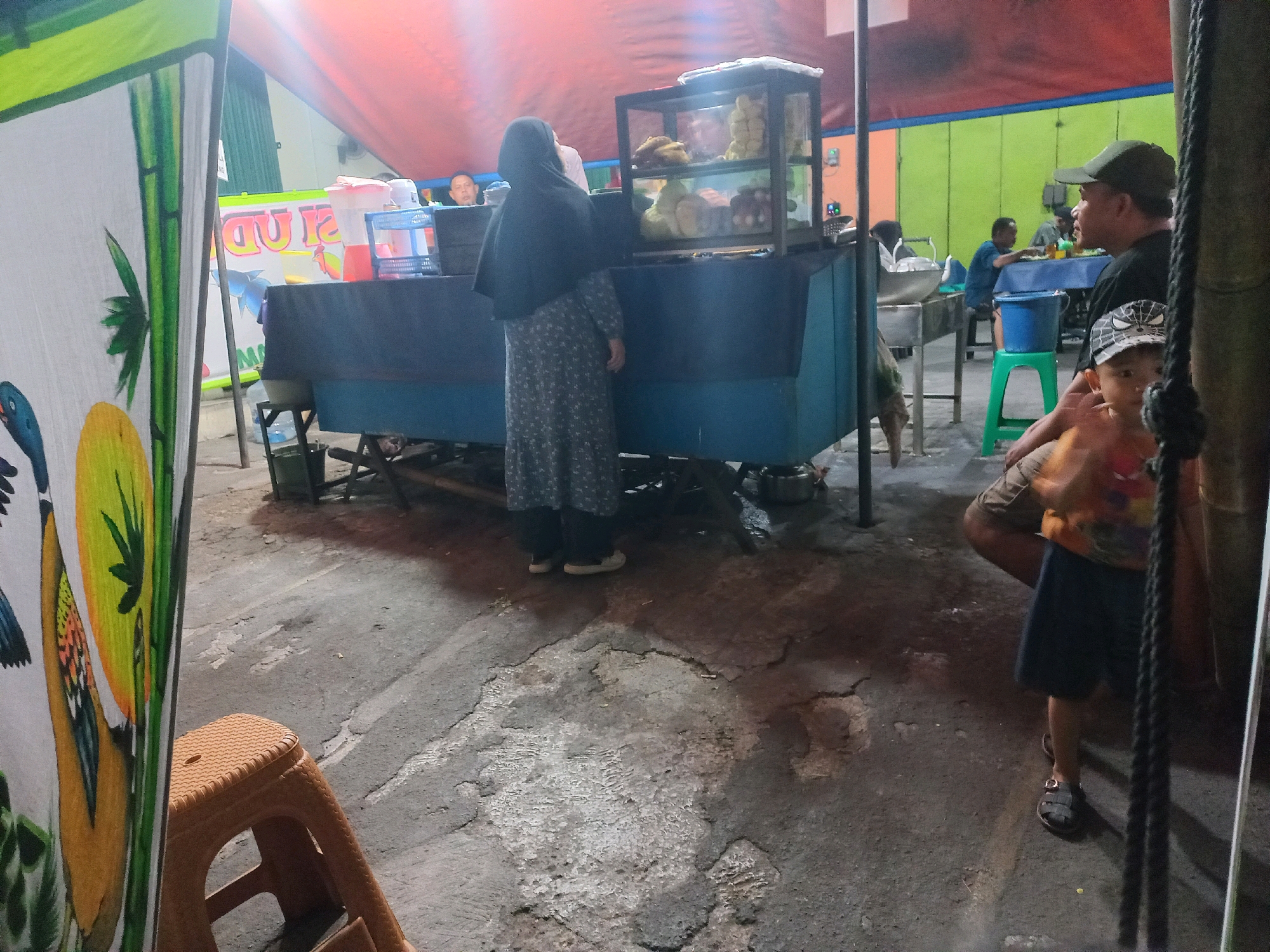 An outdoor market scene in Depok, Indonesia, is captured at dusk or night. In the foreground, a white banner with a painted image of birds and bamboo partially obscures the view of a plastic orange stool. The main area of the market features a food stall with a blue counter and a glass display case filled with food items. A woman in a dark headscarf and patterned dress stands at the counter, facing the display. Behind her, a man with a cap is visible, and further back, other people are seated at tables, seemingly eating. The environment includes a red awning overhead and a green wall in the background. The ground is asphalt with some patches of lighter colored material and cracks. A child in a patterned cap and orange shirt is partially visible in the right midground, facing towards the camera.