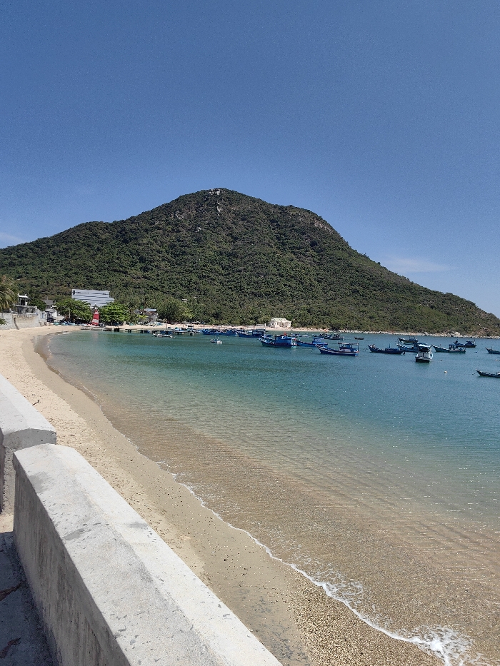A sunny coastal view showcases a sandy beach meeting clear turquoise water, filled with numerous blue boats. A large, lush green mountain rises in the background, contrasting with the bright blue sky. A grey concrete barrier runs along the edge of the beach in the foreground. Buildings and a red and white lighthouse are visible near the shore at the base of the mountain.