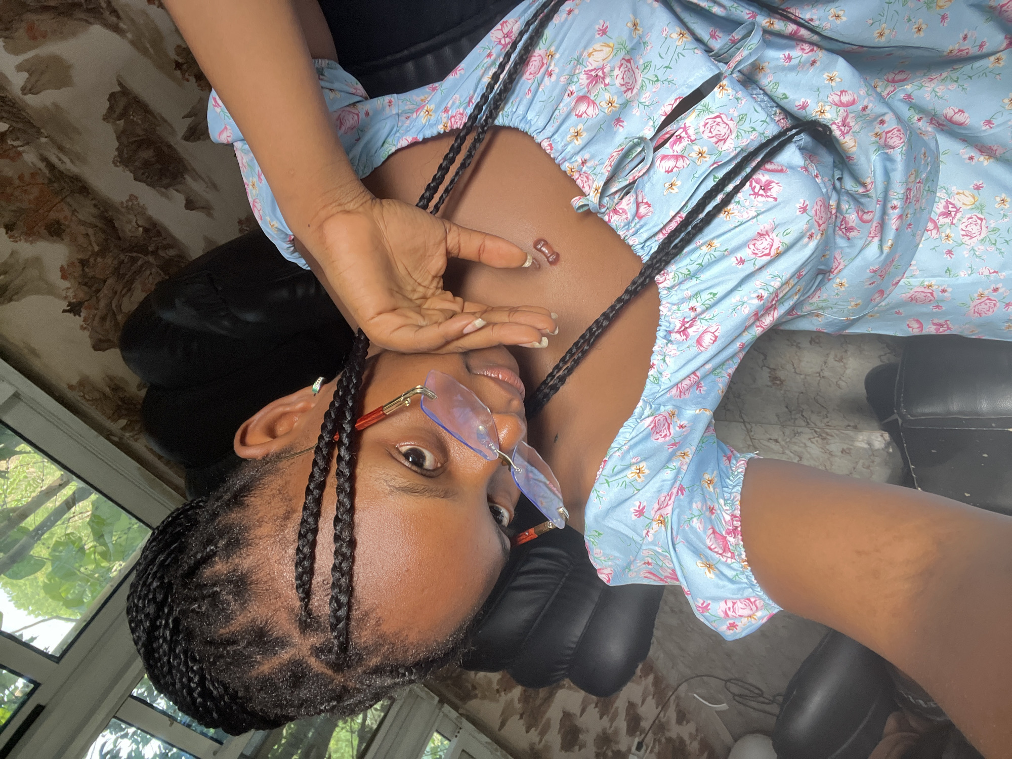 A young woman is reclining in a black leather chair. She is wearing a light blue floral print dress and clear glasses with orange frames. Her hair is braided with intricate geometric patterns at the scalp. Her left hand is raised near her face. The image appears to be taken indoors, with a window partially visible in the background. The exact location is Adankolo, Nigeria. The lighting is bright, suggesting daylight. The floor appears to be a light-colored, patterned tile.