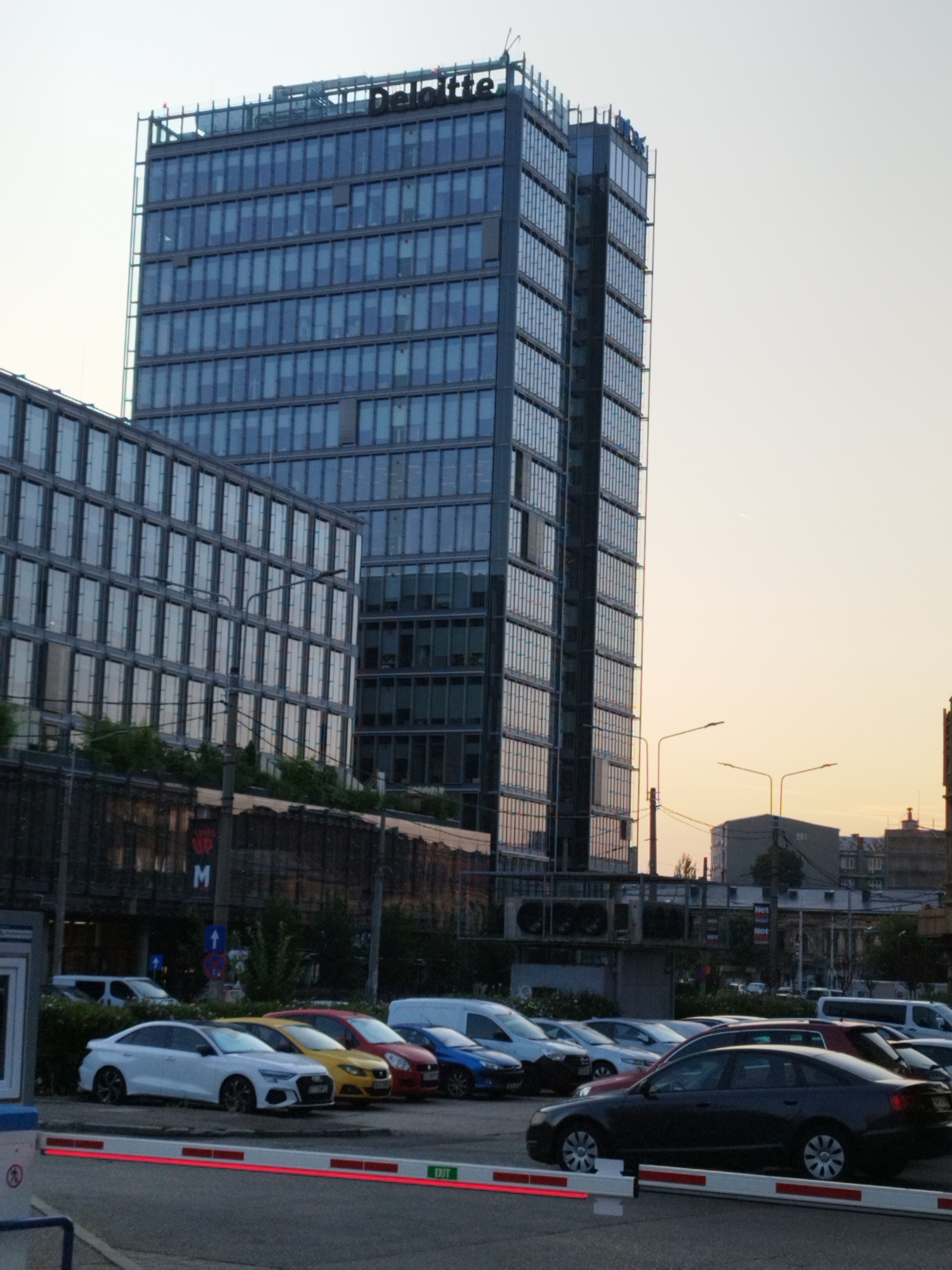 In Bucharest, Romania, a modern glass-facade skyscraper with "Deloitte" signage at its apex dominates the background, alongside another adjacent glass building. In the foreground, a parking lot is visible, with several cars parked in it, including a white sedan, a yellow sedan, a red sedan, and a blue sedan. A white van is also parked in the lot. A red and white barrier arm, likely for a parking lot entrance or exit, is positioned across the foreground, with a green "EXIT" sign on it. The sky is a pale yellow, suggesting either sunrise or sunset.