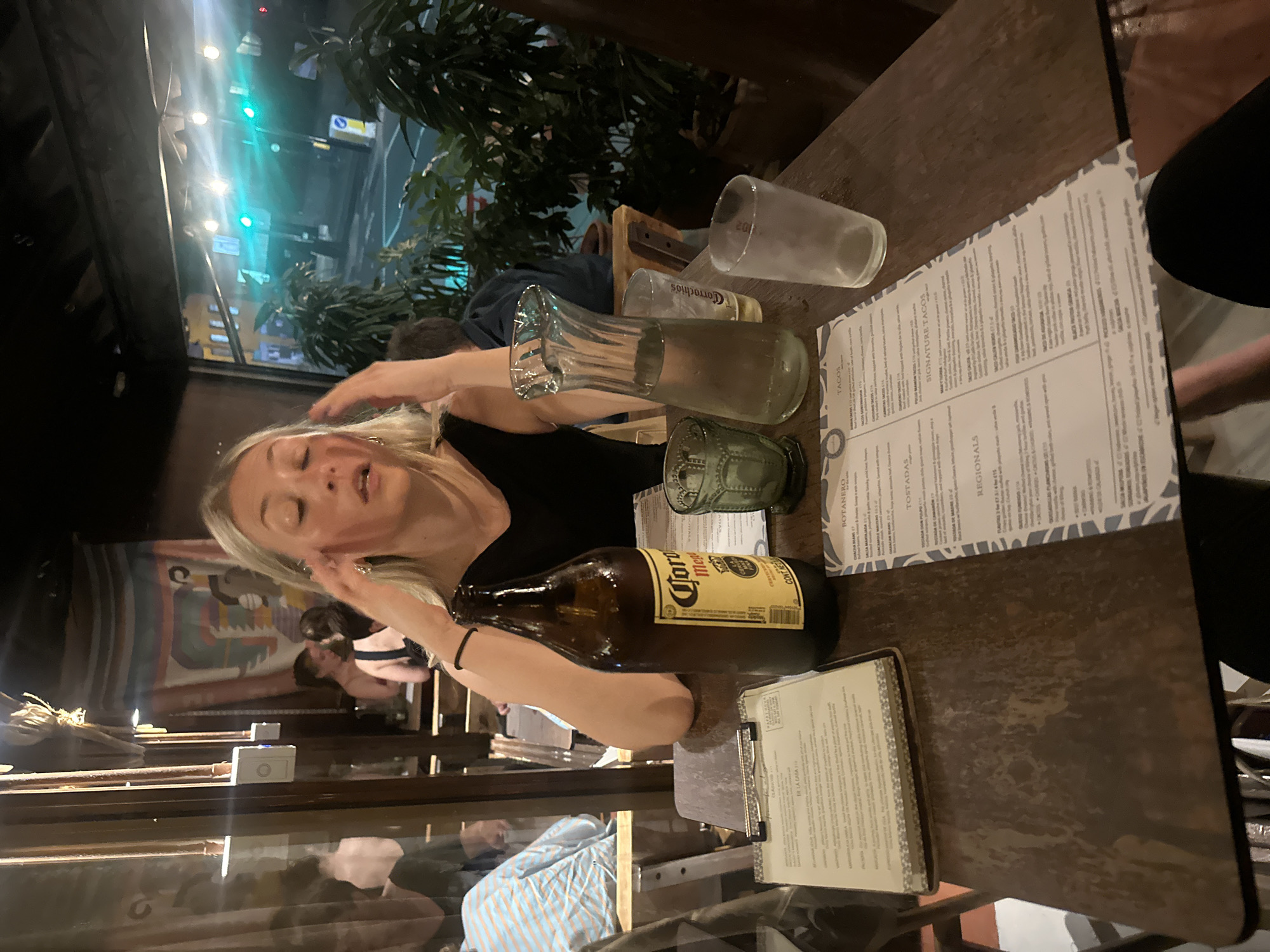 Here is a description of the image:
The image depicts a dining table scene, likely inside a restaurant or bar. A woman with blonde hair is positioned near the table, her hands near her face as if in conversation or gesturing. On the table, there are several items including a Corona beer bottle, various glassware (including what appears to be a tall glass, a shot glass, and a glass with the word "Corrachio's"), and menus. The table has a worn wooden surface, and the menus seem to have a light, patterned design.
In the background, there is a glimpse of the exterior, with some greenery and a window. Through the window, one can see what appears to be city lights and possibly a street scene, suggesting an urban setting. Additional patrons are visible seated at other tables in the background. The overall lighting is subdued, creating a warm and inviting ambiance.
It is not possible to ascertain from the image that it is specifically in the city of London, United Kingdom.